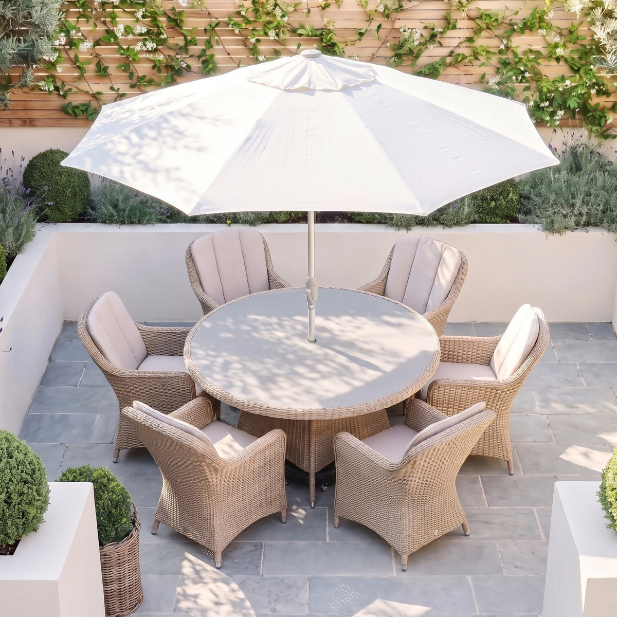 Vela 6 Seat Round Dining Set in Wheat, featuring six wicker chairs with wheat-coloured cushions and a large white umbrella, displayed on a stone patio in a garden setting.