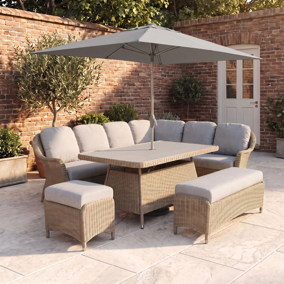 The Vela Corner Sofa Set in Wheat, paired with gray cushions, a rectangular table, umbrella, and decorative plants, creates comfortable outdoor seating on a tiled patio beside a brick wall—making it a stylish addition to your garden furniture collection.