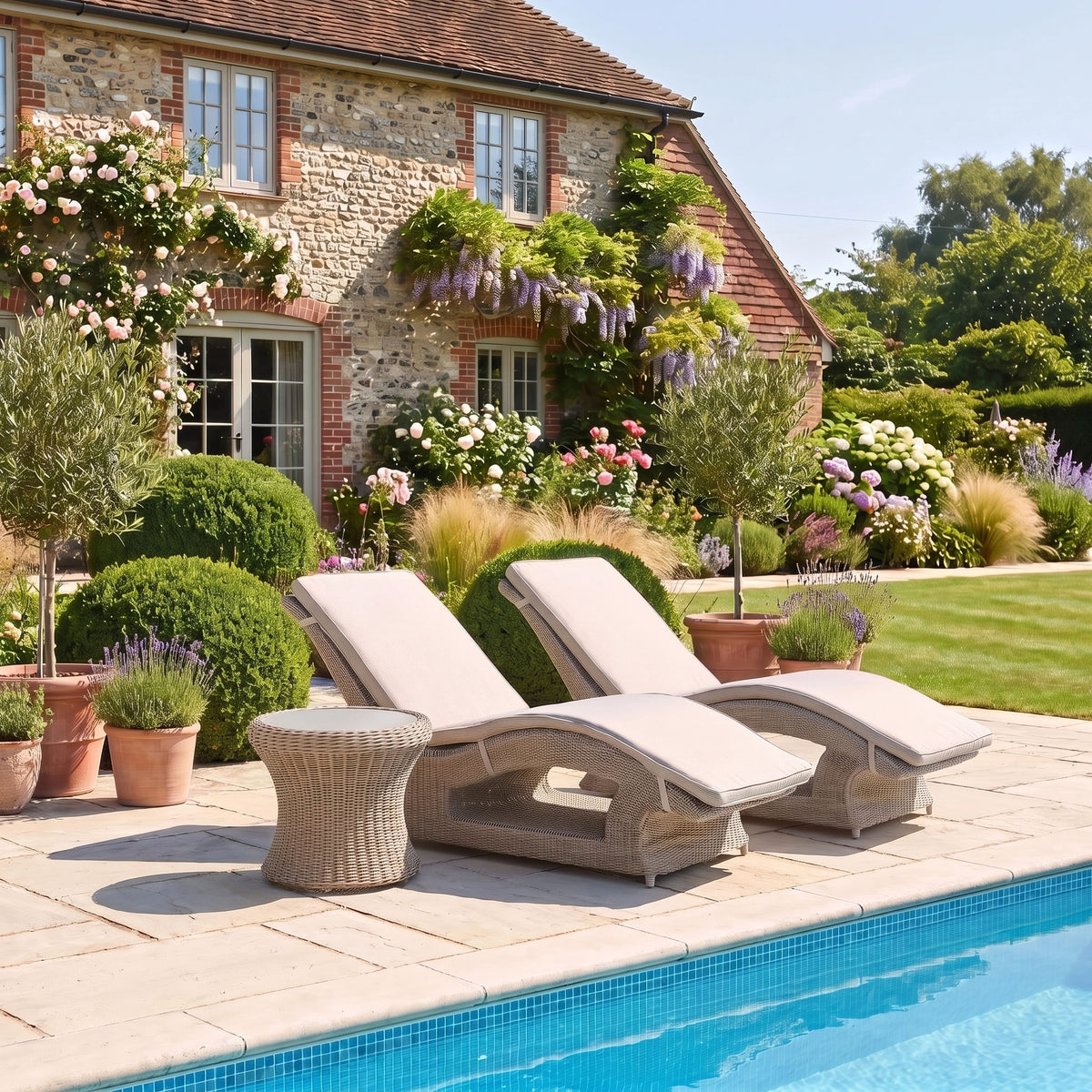 Two lounge chairs and a Vela Round Side Table in Wheat with a tempered glass top sit beside a pool, with a garden and brick house in the background.
