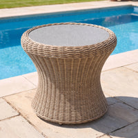 The Vela Round Side Table in Wheat, featuring a round wicker base and a sleek stone top, sits on a patio by the swimming pool—making it the perfect outdoor side table for relaxing in style.