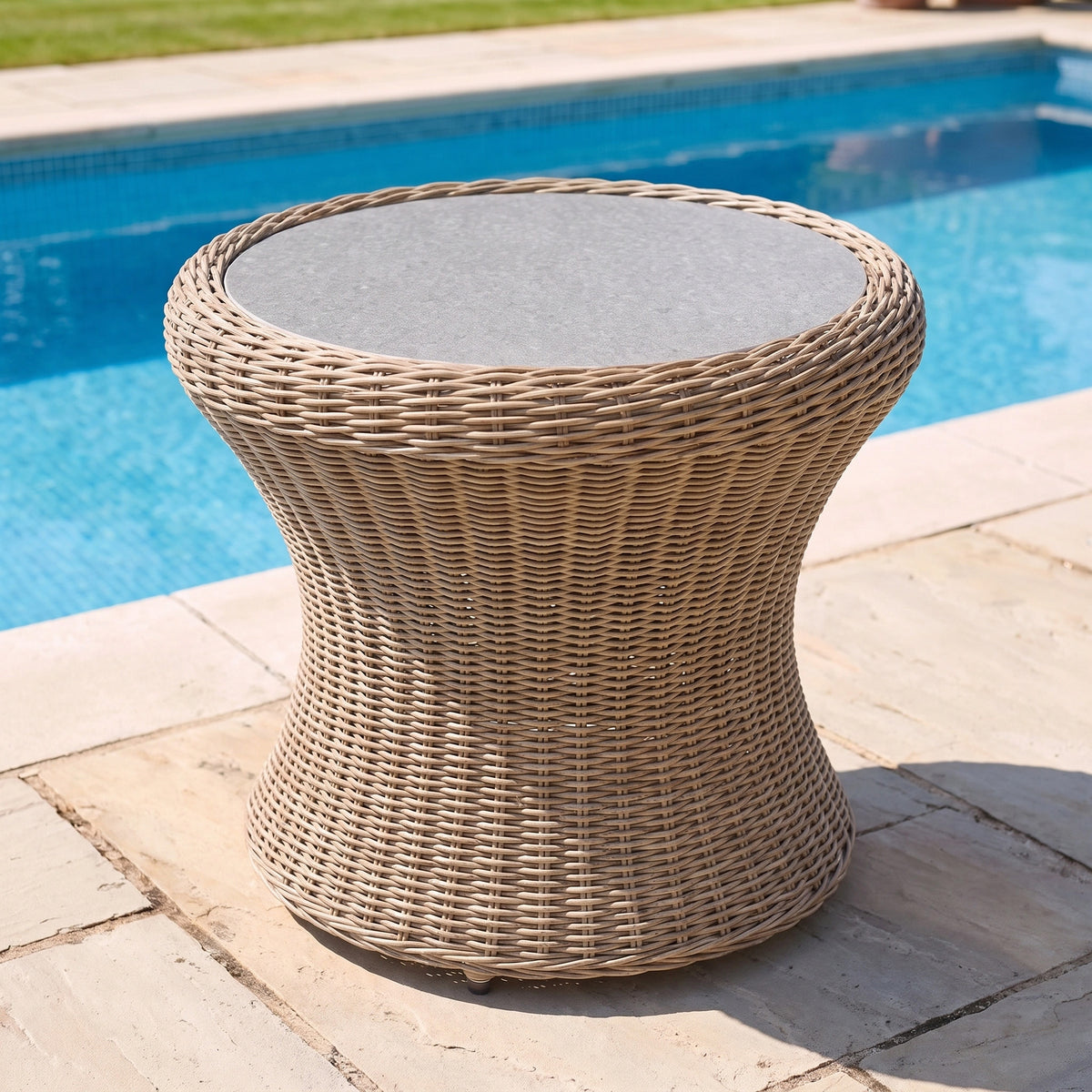 The Vela Round Side Table in Wheat, featuring a round wicker base and a sleek stone top, sits on a patio by the swimming pool—making it the perfect outdoor side table for relaxing in style.