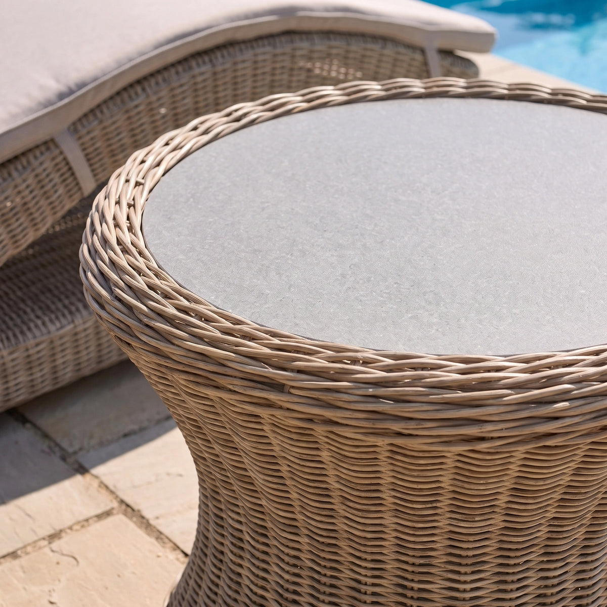 The Vela Round Side Table in Wheat, featuring a tempered glass top, sits round and inviting by the pool, perfectly complemented by a matching wicker lounge chair and stone patio for an ideal outdoor side table arrangement.