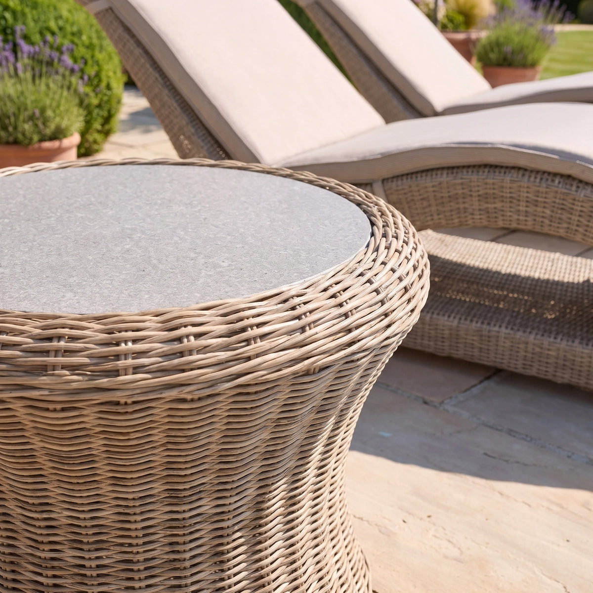 The Vela Round Side Table in Wheat features a wicker design with a stone top, making it the perfect outdoor side table to place beside cushioned wicker loungers on a sunny patio.