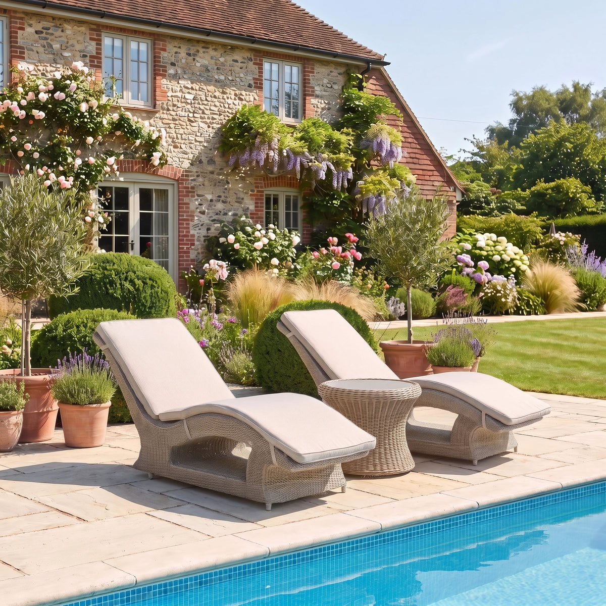 Two Vela Curved Sun Loungers in Wheat sit by a tranquil pool, with a stone house and blooming garden in the background.