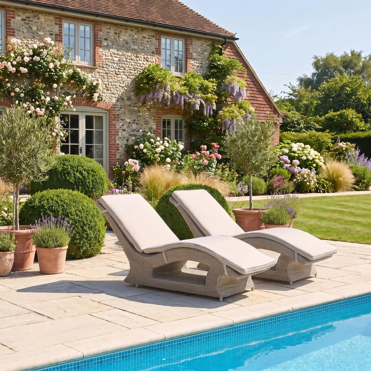 Two Vela Curved Sun Loungers in Wheat sit by the pool, with a lush garden and charming stone house in the background.