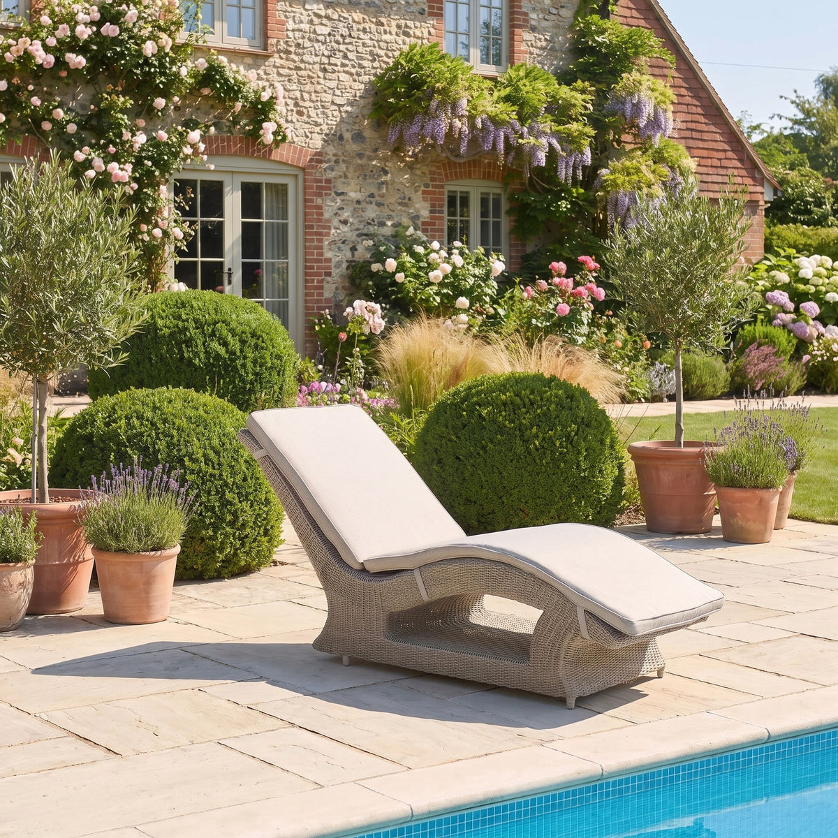 A Vela Curved Sun Lounger in Wheat sits beside the pool, surrounded by potted plants and a lush garden in front of a house—this inviting outdoor scene is enhanced by the stylish sun lounger, perfect for relaxing under the sun.