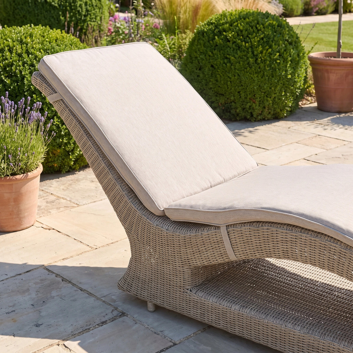 Vela Curved Sun Lounger in Wheat with a beige cushion on a stone patio, surrounded by potted plants and greenery—this inviting outdoor lounger blends comfort and style for the perfect relaxation spot.