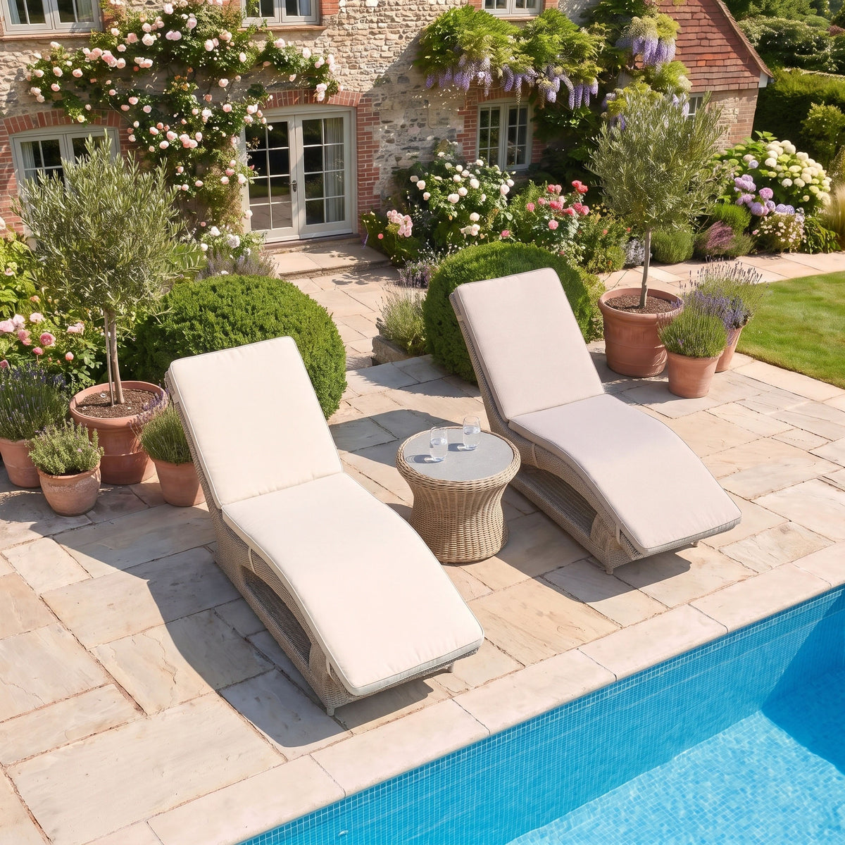 Two Vela Curved Sun Loungers in Wheat and a small table by a pool, surrounded by potted plants and a stone house.