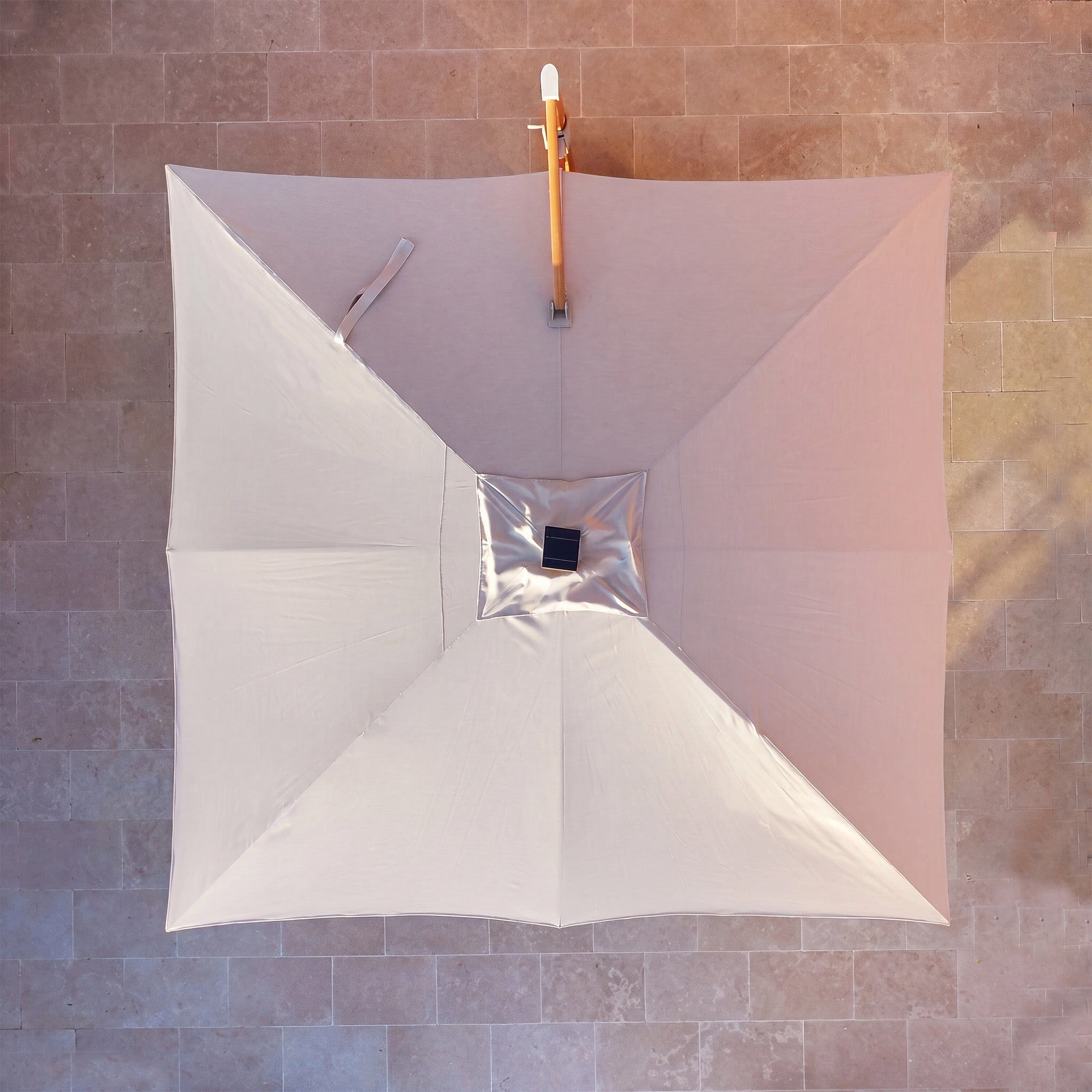 The Volos 3m x 3m Square Cantilever Parasol with LED Lighting and 120kg Granite Base in Wheat and Wood Effect is viewed from above, its UV resistant canopy casting a shadow on stone tiles.