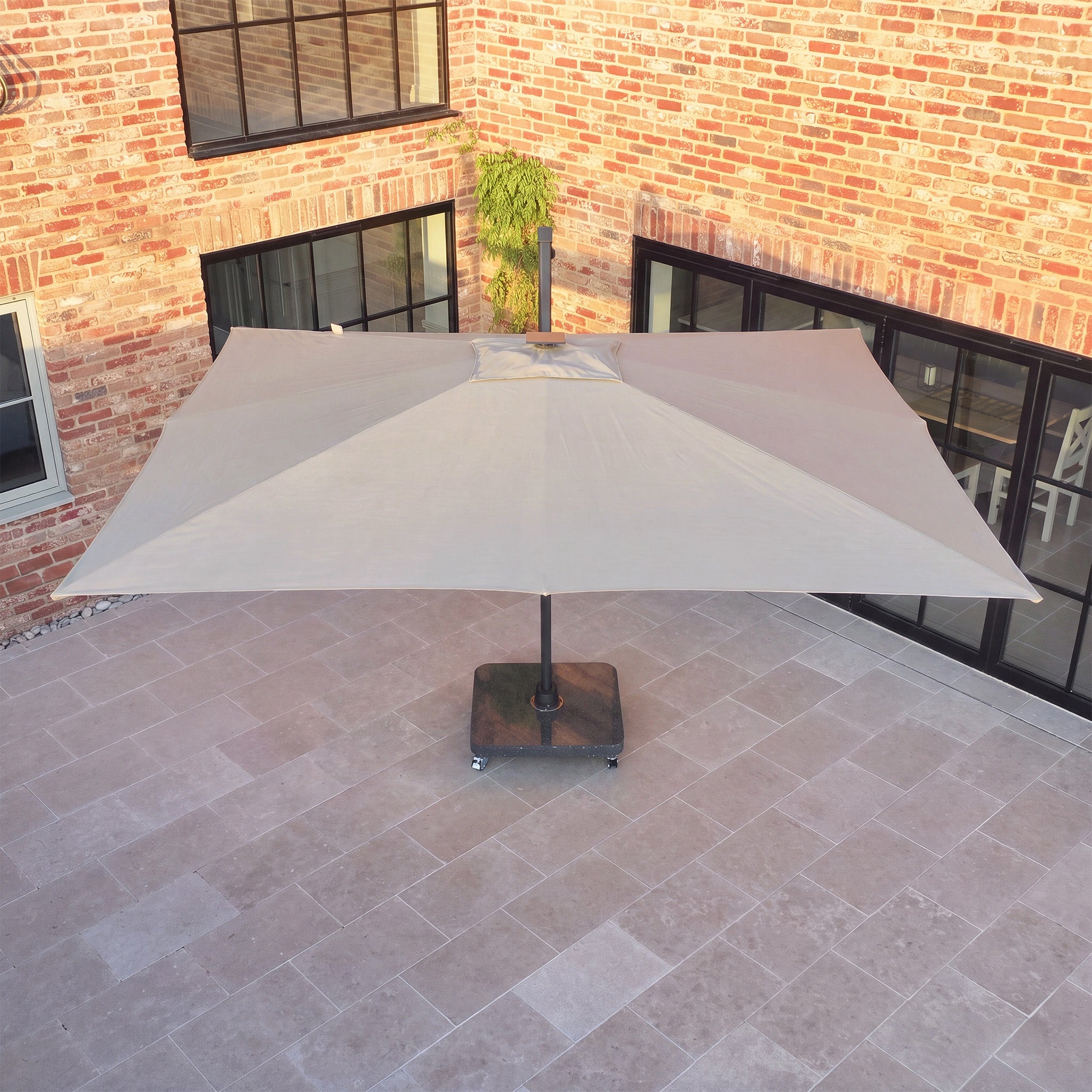 A Volos 3m x 4m Aluminium Cantilever Parasol with LED Lighting and a 120kg granite base in Wheat stands on a tiled courtyard beside a brick building with large windows.