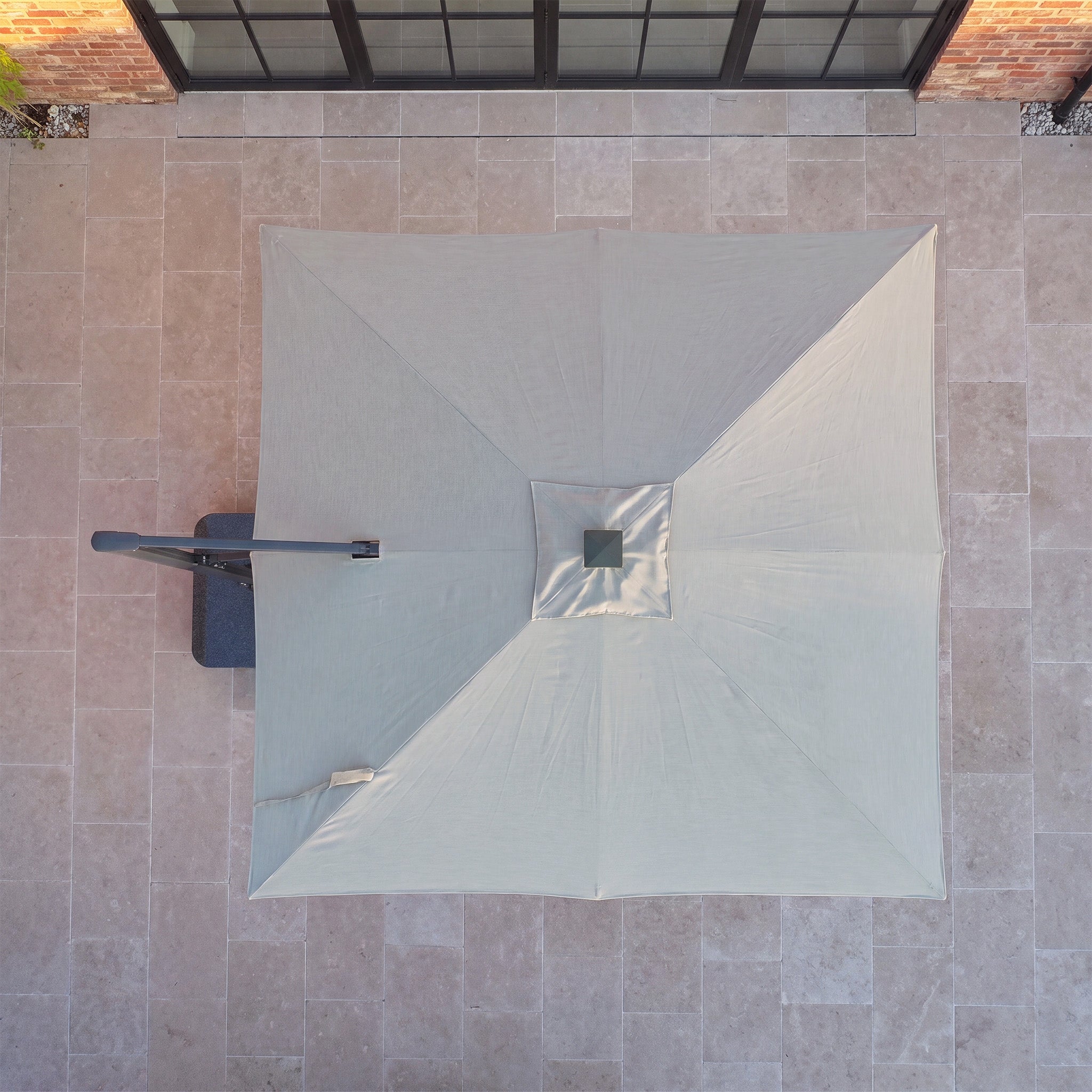 The Volos 3m x 3m Square Cantilever Parasol with LED Lighting and 120kg Granite Base in Wheat is seen from above, positioned on light stone tiles near a building with glass doors.