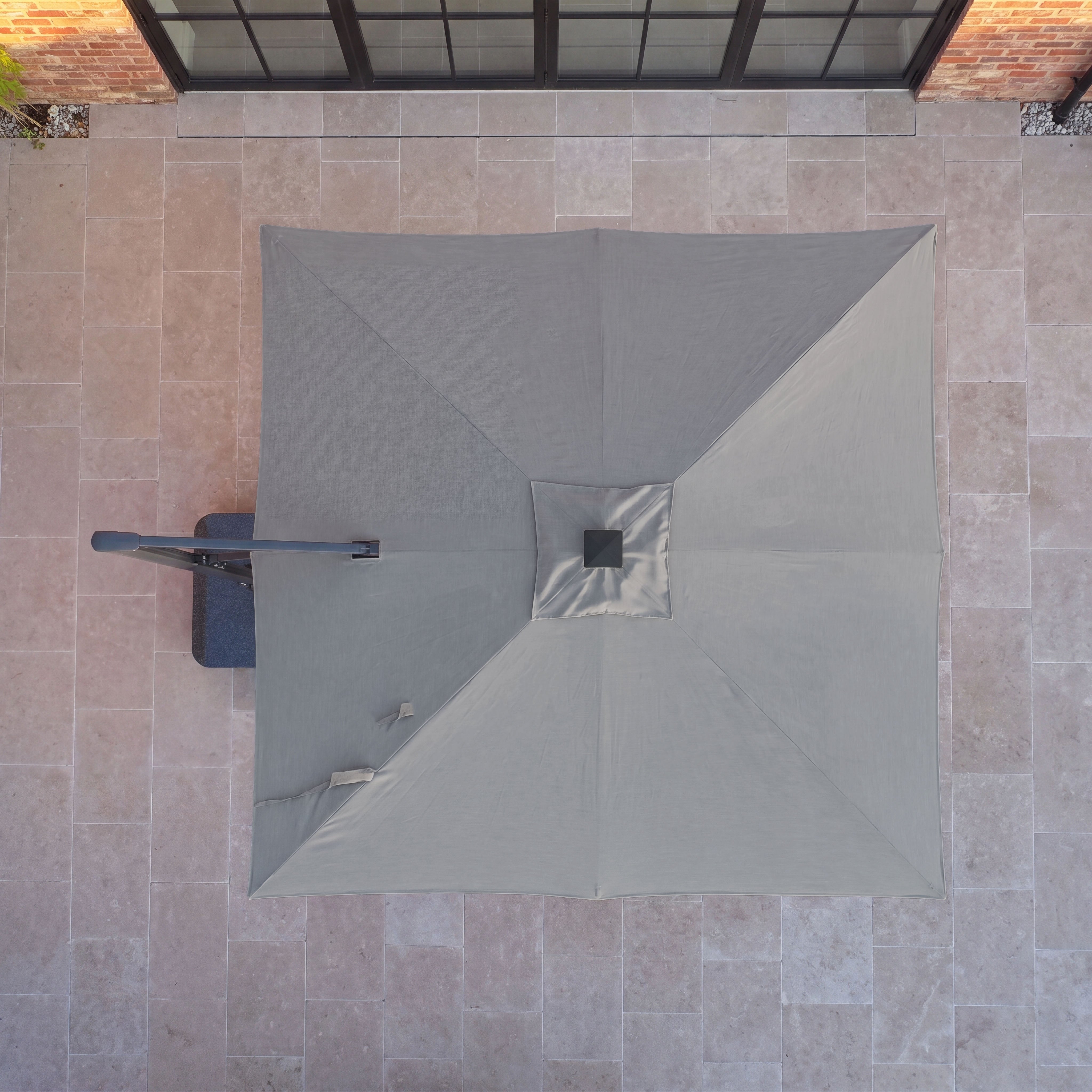 The Volos 3m x 3m Square Cantilever Parasol with LED Lighting and a 120kg granite base in Slate, featuring UV resistance, is shown from above on a tiled patio next to glass doors and a brick wall.
