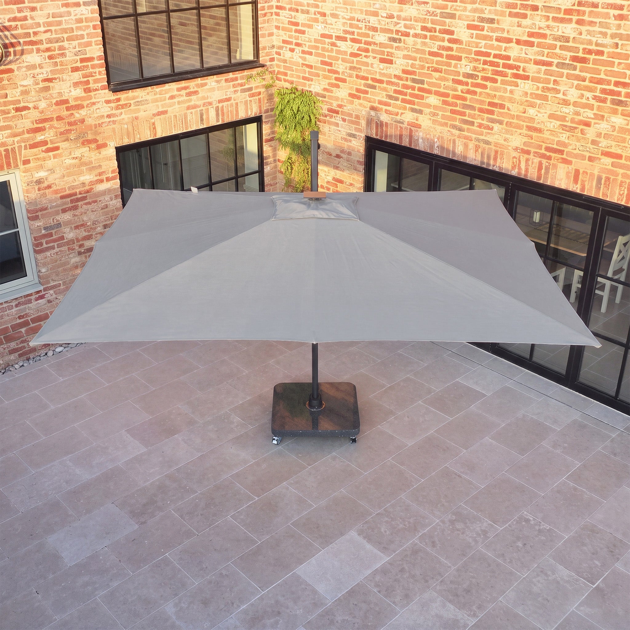 The Volos 3m x 4m Rectangular Cantilever Parasol with LED Lighting and a 120kg granite base in slate stands on the tiled courtyard, its UV resistant canopy open, framed by brick walls and glass doors in the background.