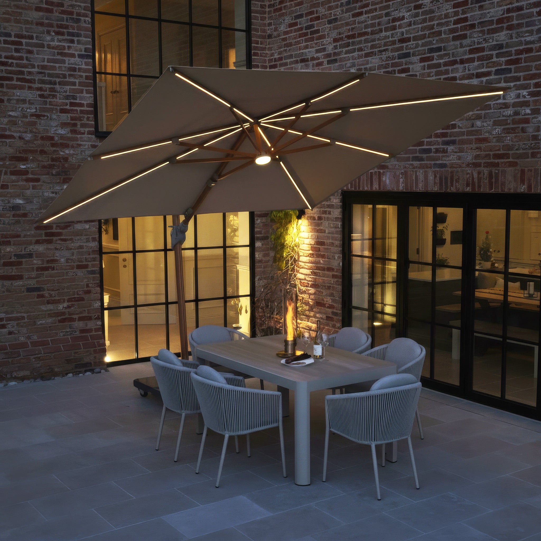 A modern patio at dusk features the Volos 3m x 3m Square Cantilever Parasol with LED Lighting and 120kg granite base in wheat and wood effect, alongside a dining table and chairs against a brick building.