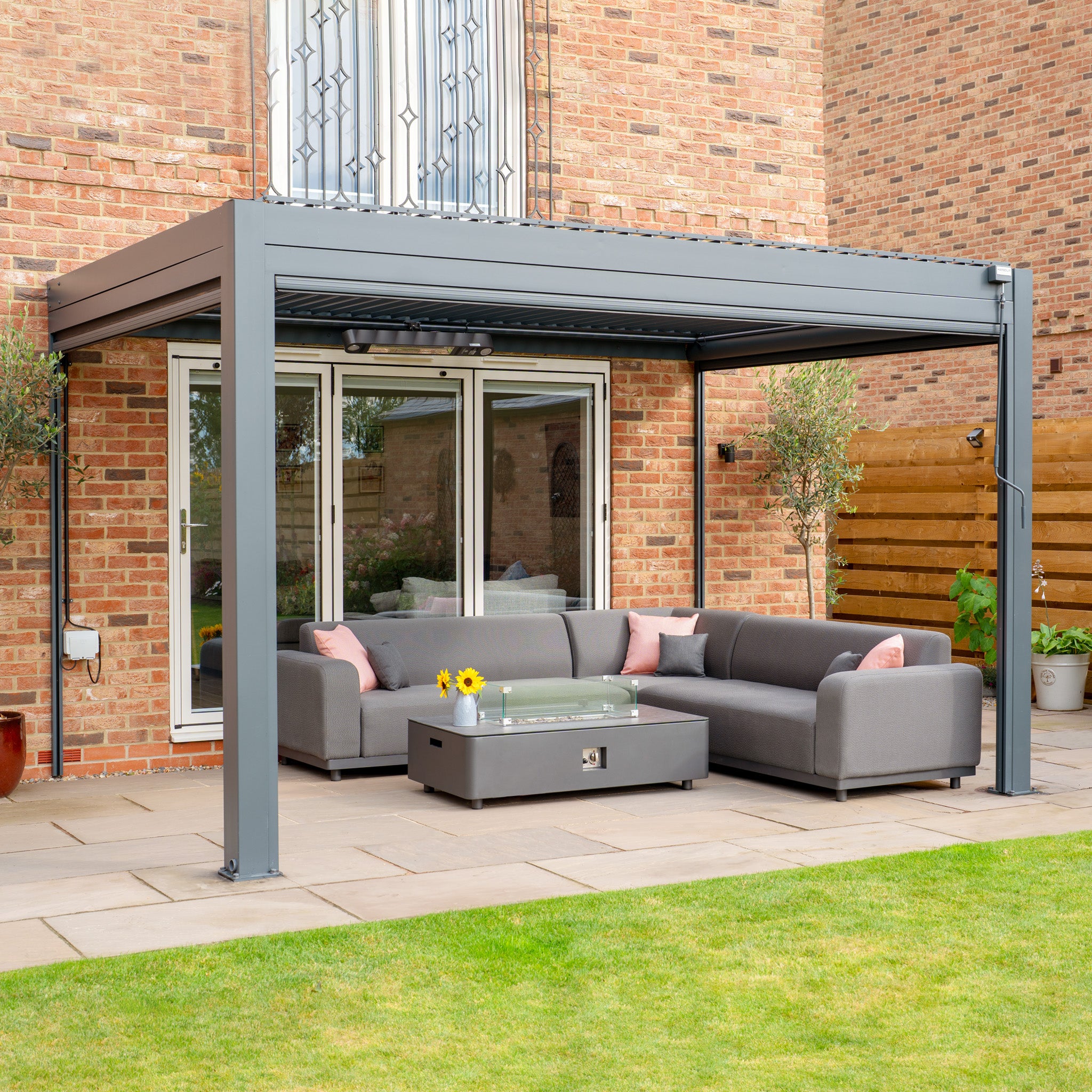 The modern patio boasts a gray sectional sofa, glass-top table, and a stylish PergoSTET Plus pergola with LED lighting and waterproof roof. Bright sunflowers and vibrant cushions enhance the setup beautifully.