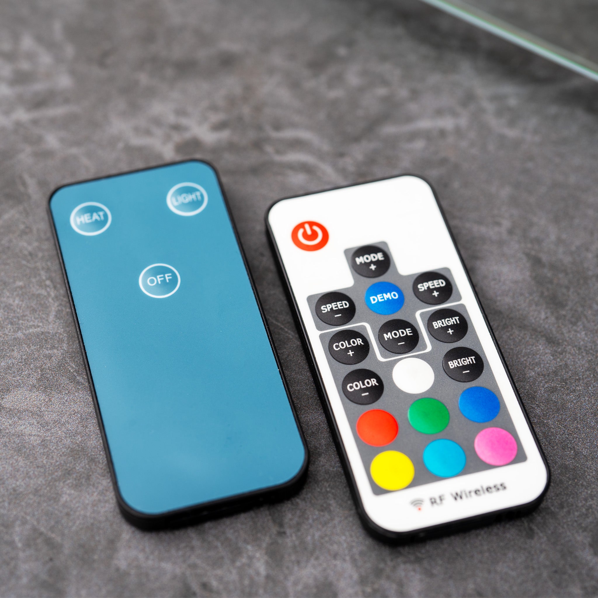 Two remote controls on a gray surface; one blue with three buttons, the other white with multiple colorful buttons, perfect for adjusting your PergoSTET Plus 3m x 4m Rectangular Wall Mounted Pergola with 3 Drop Sides and LED Lighting in Grey.
