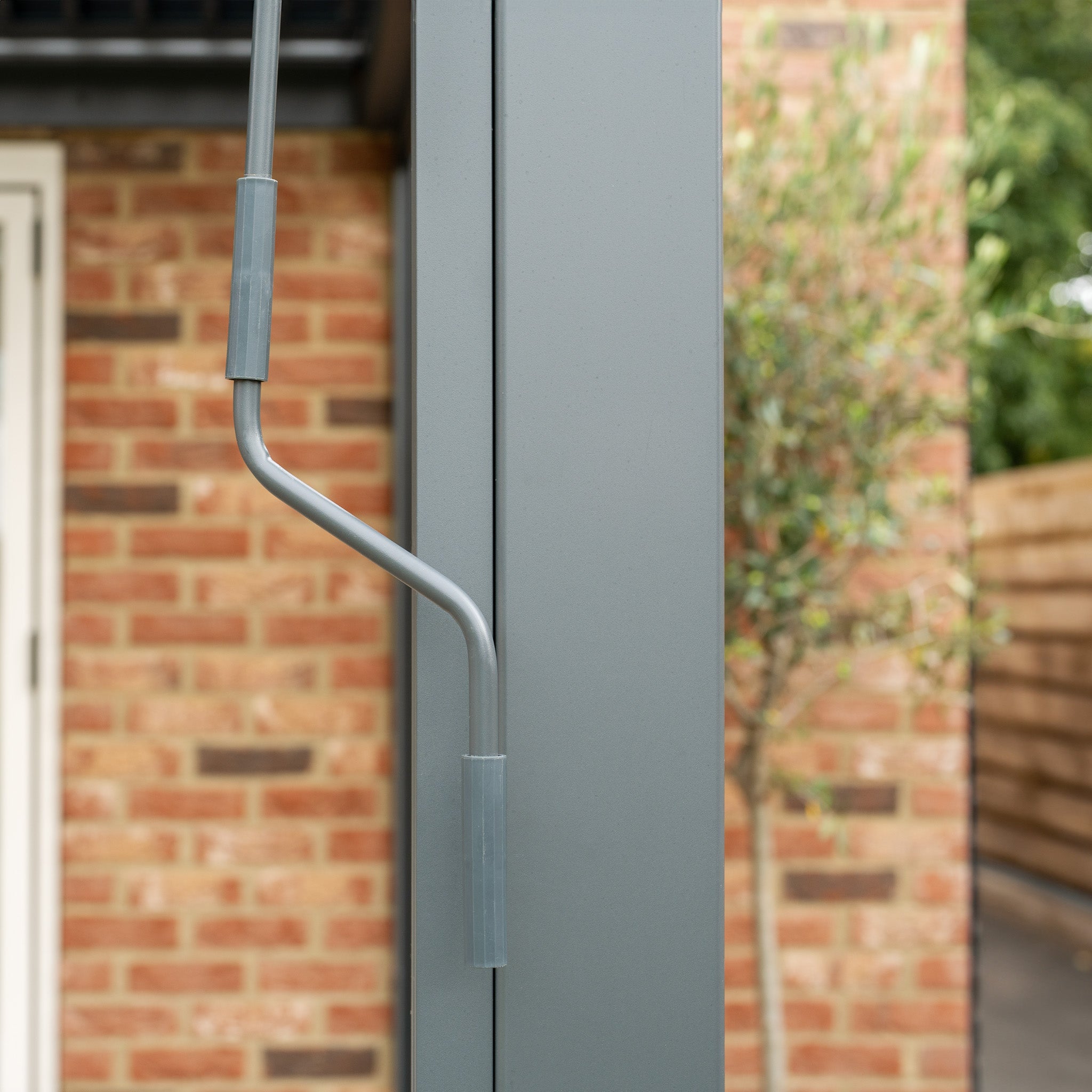 Close-up of a metal turn handle on a gray pole, set against a brick wall and greenery. Ideal for the PergoSTET Plus 3m x 4m Rectangular Wall Mounted Pergola with 3 Drop Sides and LED Lighting in Grey.
