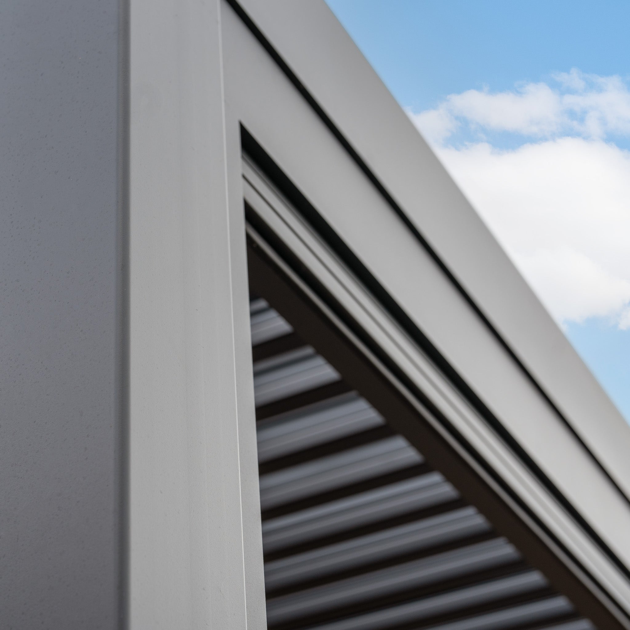 Close-up of the PergoSTET Plus 3m x 3m Square Wall Mounted Pergola in Grey, featuring parallel slats and tilting louvres, set against a blue sky with white clouds.