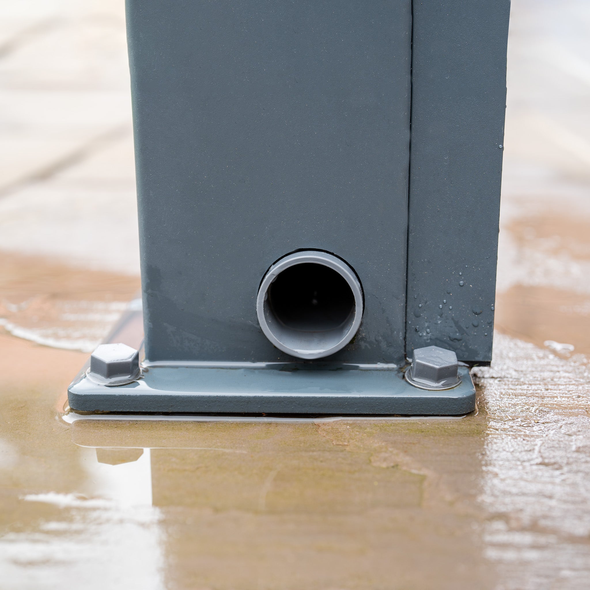 A close-up of the PergoSTET Plus Pergola shows a gray metal structure with a circular drainage pipe and bolts on wet concrete. Its sleek design mirrors modern Pergolas, offering an intriguing visual play while enhancing functionality, complete with 3 drop sides and LED lighting.