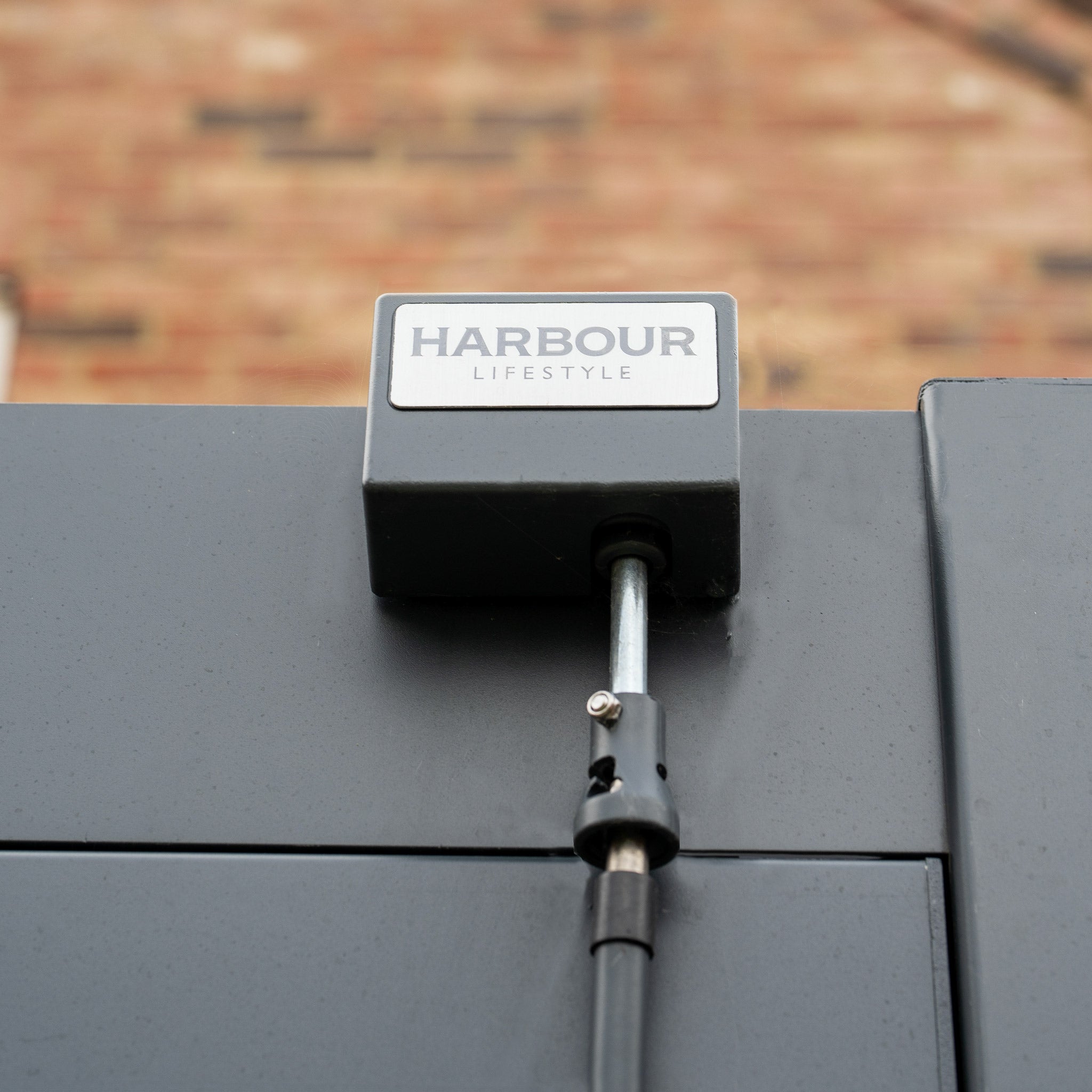 Close-up of a "Harbour Lifestyle" PergoSTET Plus 3m x 3m Square Pergola with 3 drop sides and LED lighting in grey, mounted on a pole against a brick wall, highlighting its sleek design.