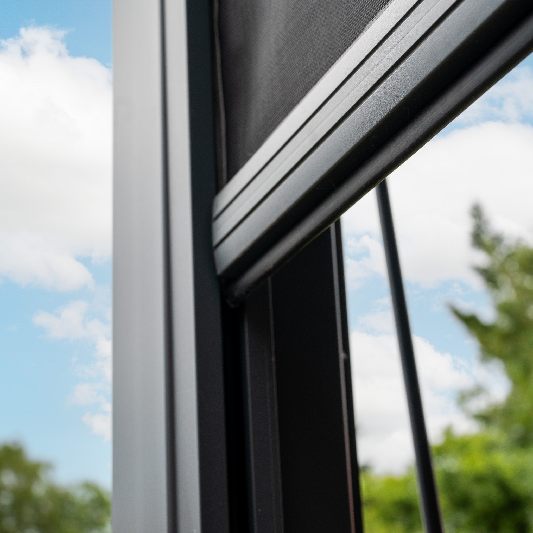 A window with a sliding screen reveals a partly cloudy sky and tree tops under the shadow of the sleek PergoSTET Plus Rectangular Wall Mounted Pergola, featuring 3 drop sides and LED lighting in Grey.