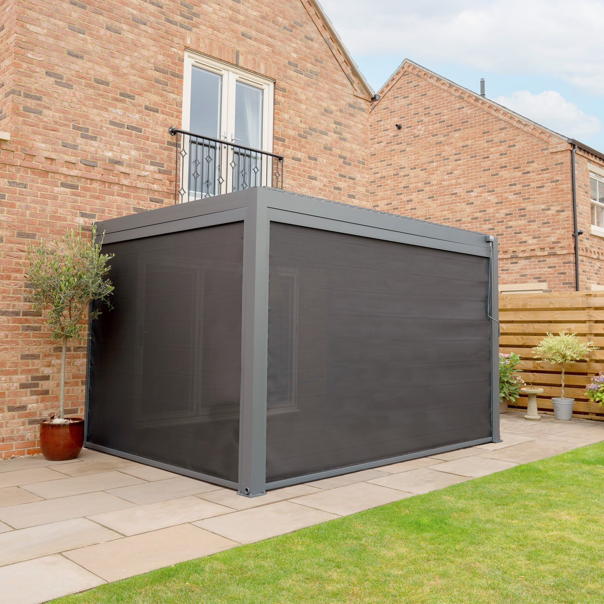 The PergoSTET Plus pergola, a wall-mounted 3m x 4m design with LED lighting and three drop sides in grey, features a waterproof roof seamlessly attached to a brick house. It sits on a patio surrounded by lush plants.