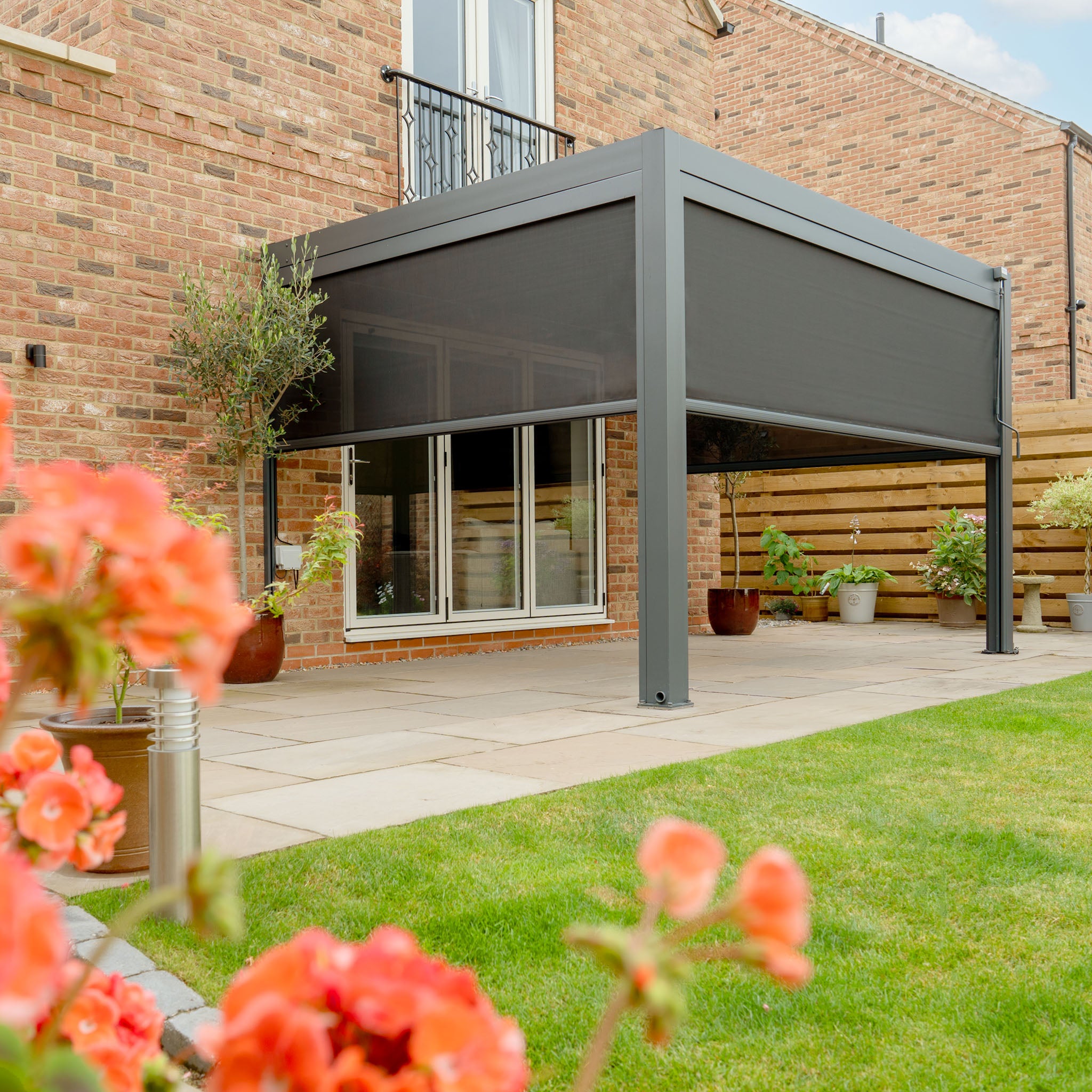 A modern patio with a sleek PergoSTET Plus 3m x 4m Rectangular Wall Mounted Pergola featuring 3 drop sides and LED lighting in grey, accented by stylish sunshades and potted plants against a brick house, with vibrant flowers flourishing in the foreground.