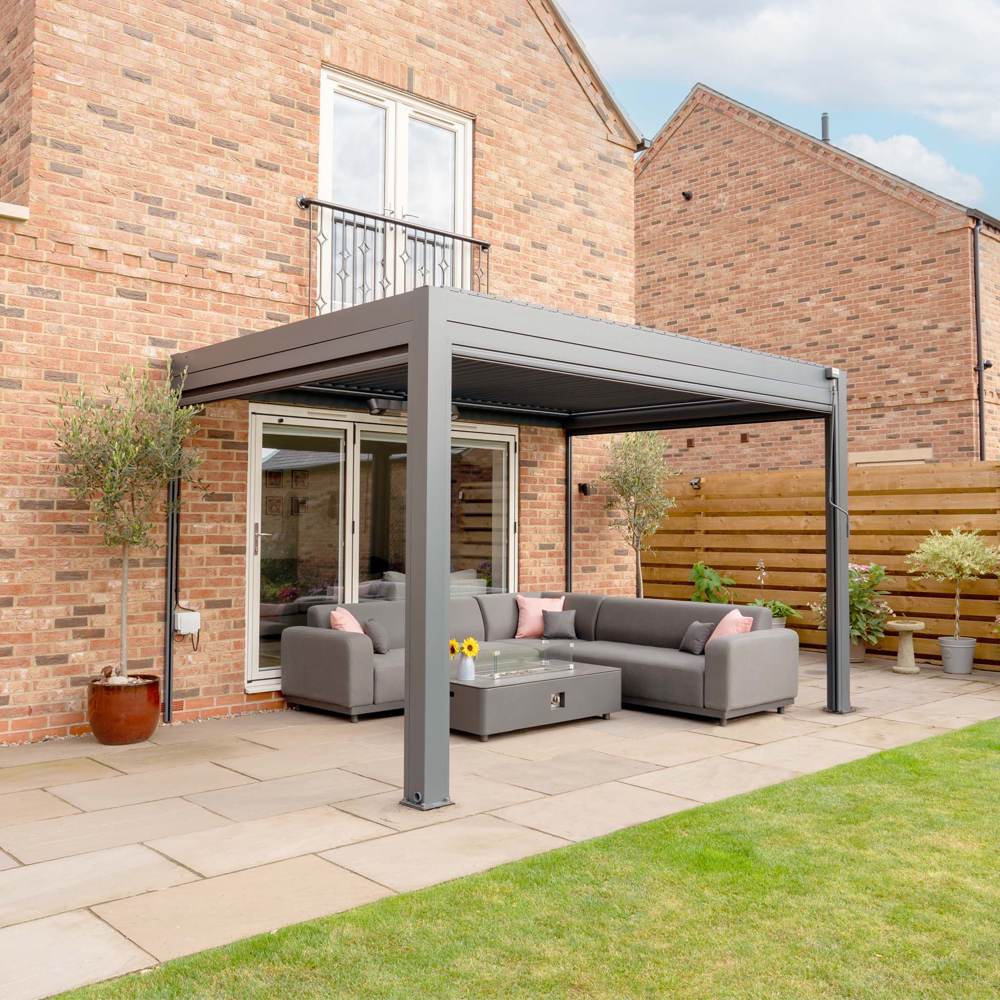 A modern patio features a PergoSTET Plus 3m x 4m gray pergola with three drop sides and LED lighting, a sectional sofa, and a glass-topped table; brick house and wooden fence are in the background.