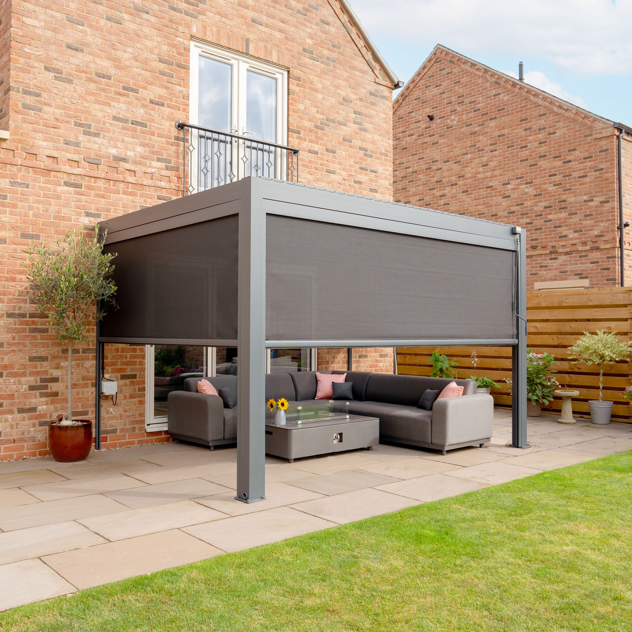 Contemporary outdoor seating area featuring gray sofas beneath a PergoSTET Plus 3m x 4m Rectangular Wall Mounted Pergola with 3 Drop Sides and LED Lighting in Grey, adjacent to a brick house on a paved patio.