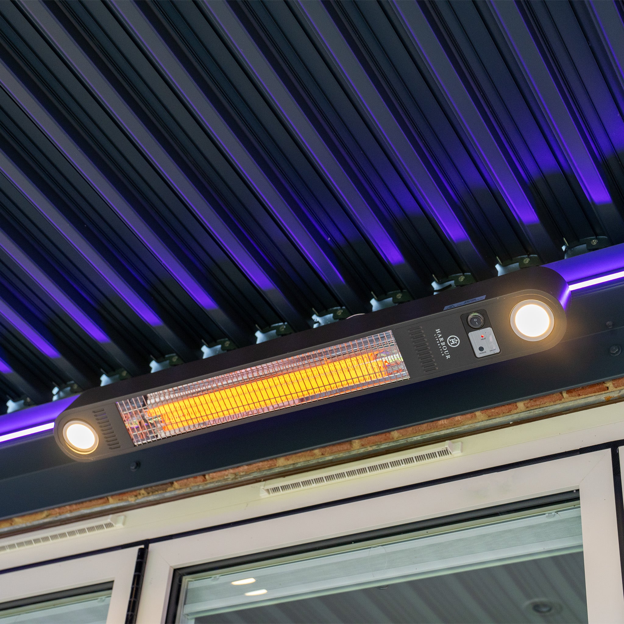 The ceiling-mounted heater with glowing element and two circular lights under a dark ribbed ceiling enhances the modern aesthetic of the PergoSTET Plus 3m x 3m Square Wall Mounted Pergola with 3 Drop Sides and LED Lighting in Grey.
