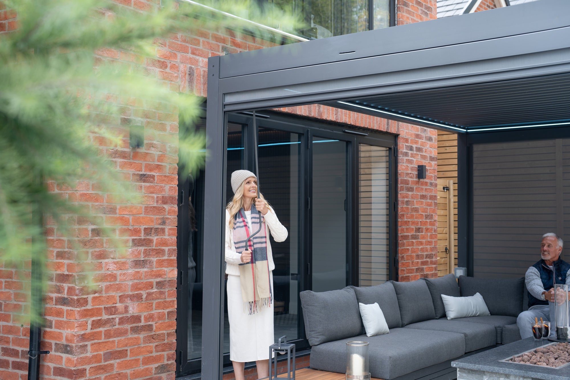 A lifestyle of image of a 3m x 4m Pergostet Plus Pergola in grey. The customer is using the hand crank to adjust the louvres of the pergola.