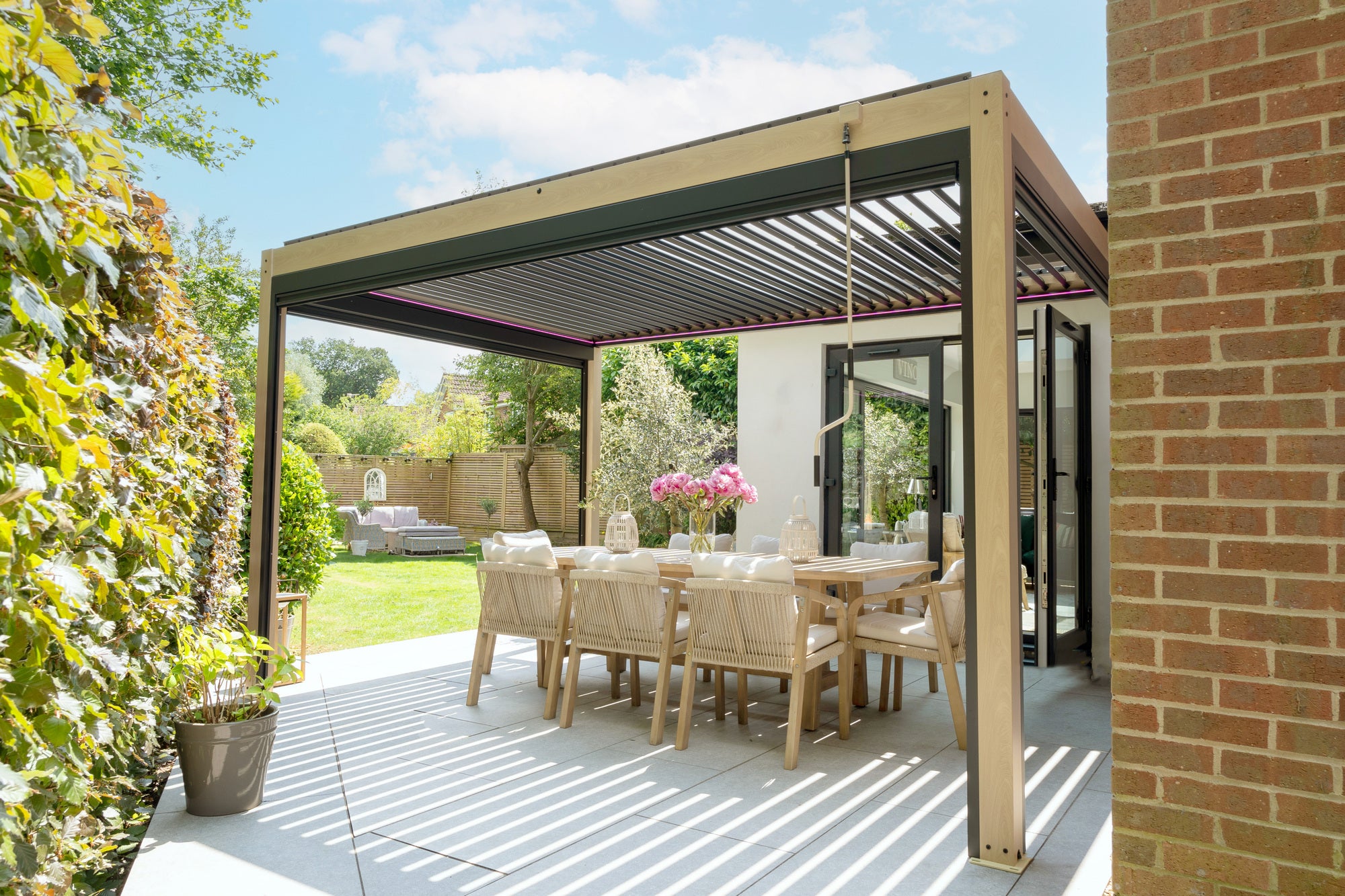 A modern outdoor dining area with a wooden pergola, table, chairs, and a vase of flowers. The area is adjacent to a brick building and overlooks a green garden with a hedge.