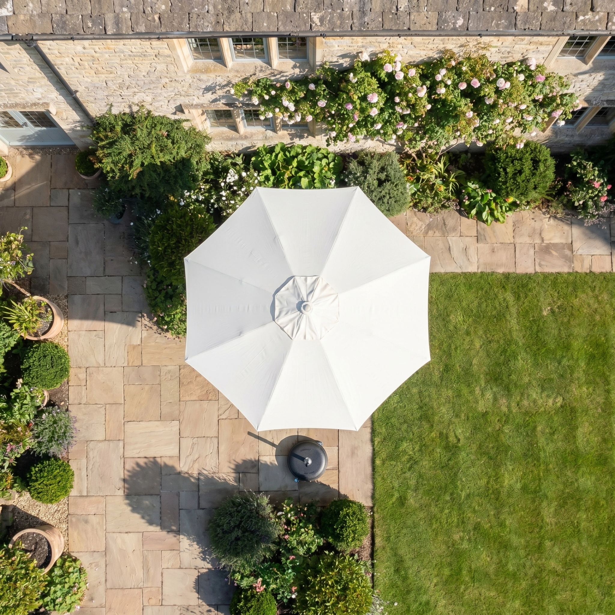 Overhead view of a patio featuring the Zelos 2.5M Round Parasol with LED Lighting in Cream for outdoor sun protection, surrounded by potted plants and a stone house adorned with climbing roses.