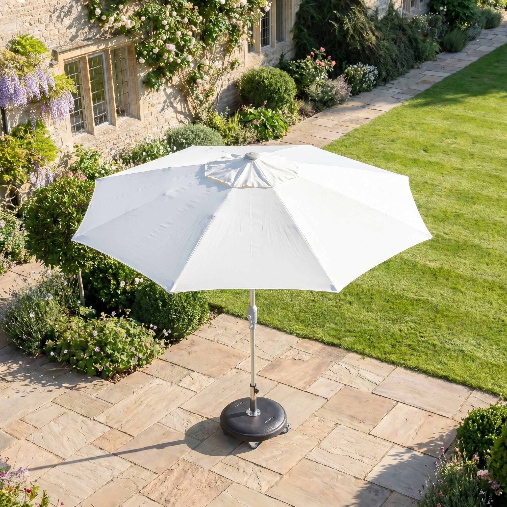 The Zelos 2.5M Round Parasol with LED Lighting in Cream offers excellent sun protection on a stone terrace beside a house, perfect for enjoying your well-maintained garden in comfort and style.