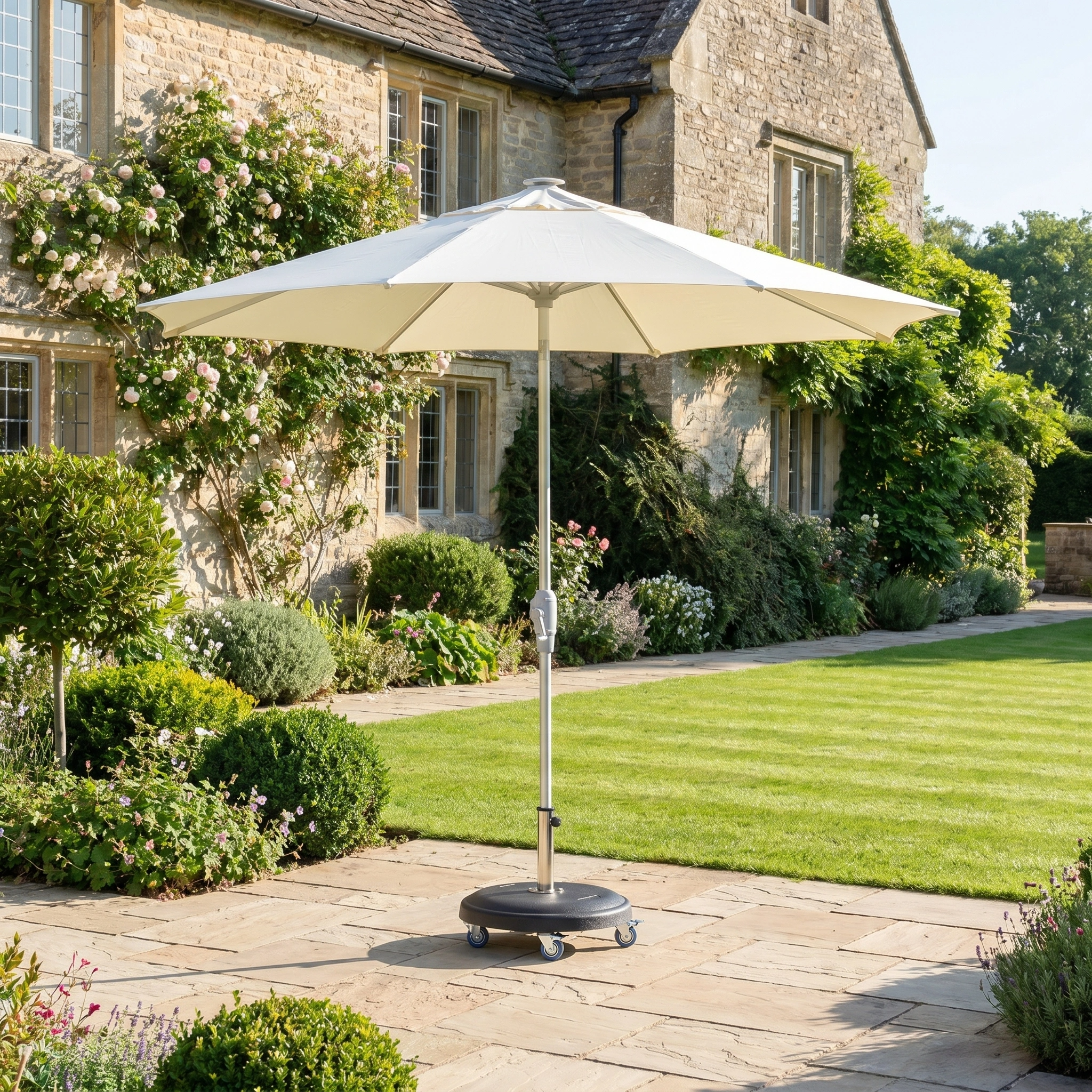The Zelos 2.5M Round Parasol with LED Lighting in Cream is positioned on a stone patio in a landscaped garden next to a rustic stone house.