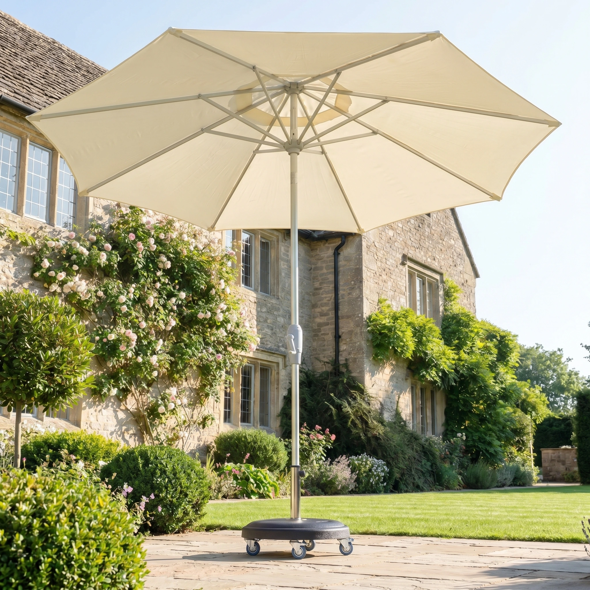 The Zelos 2.5M Round Parasol with LED Lighting in Cream stands on a stone patio by a stone house with climbing plants, offering stylish sun protection for your garden.