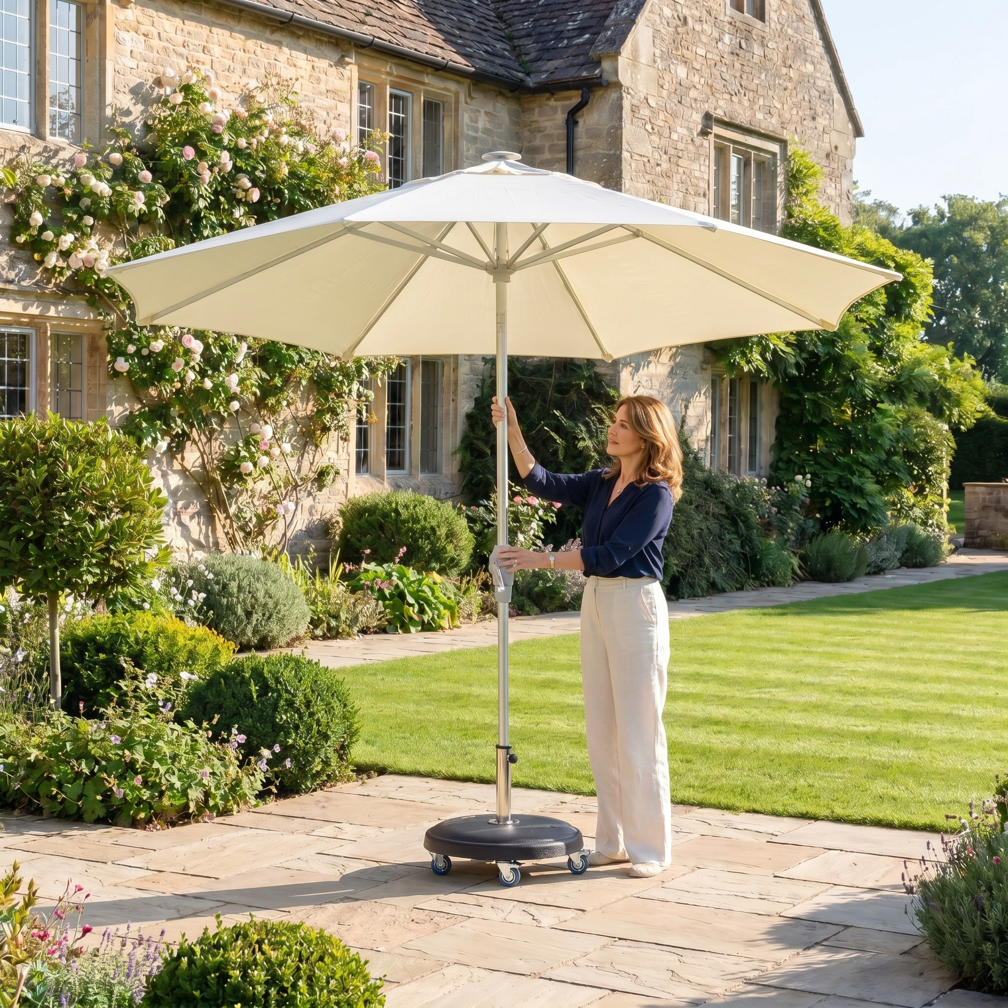 A woman adjusts the Zelos 2.5M Round Parasol with LED Lighting in Cream on a sunny patio beside a stone house, enjoying stylish shade and illuminated outdoor sun protection.