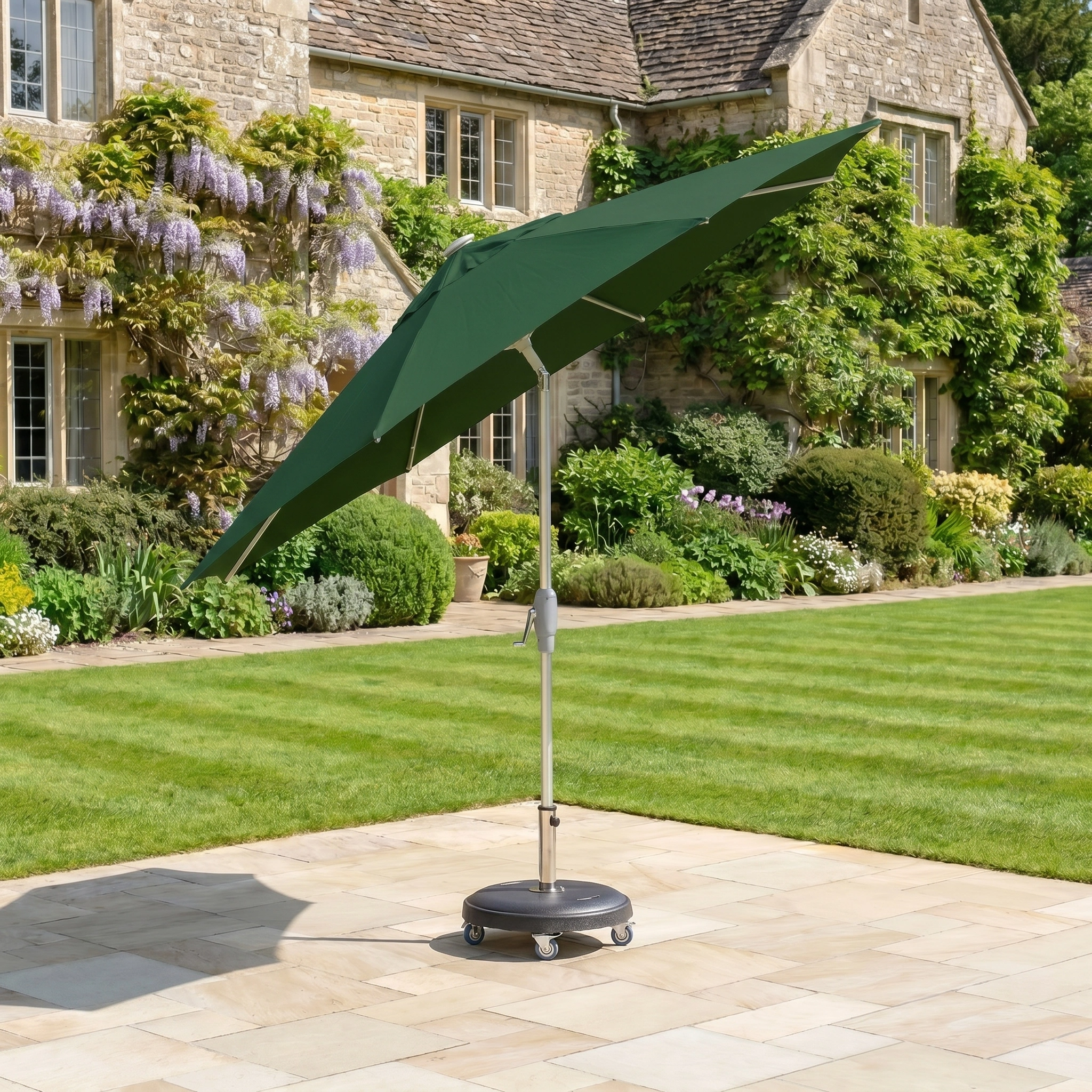 The Zelos 2.5M Round Parasol with LED Lighting in Green stands on a stone terrace in a garden, with a large house visible in the background.