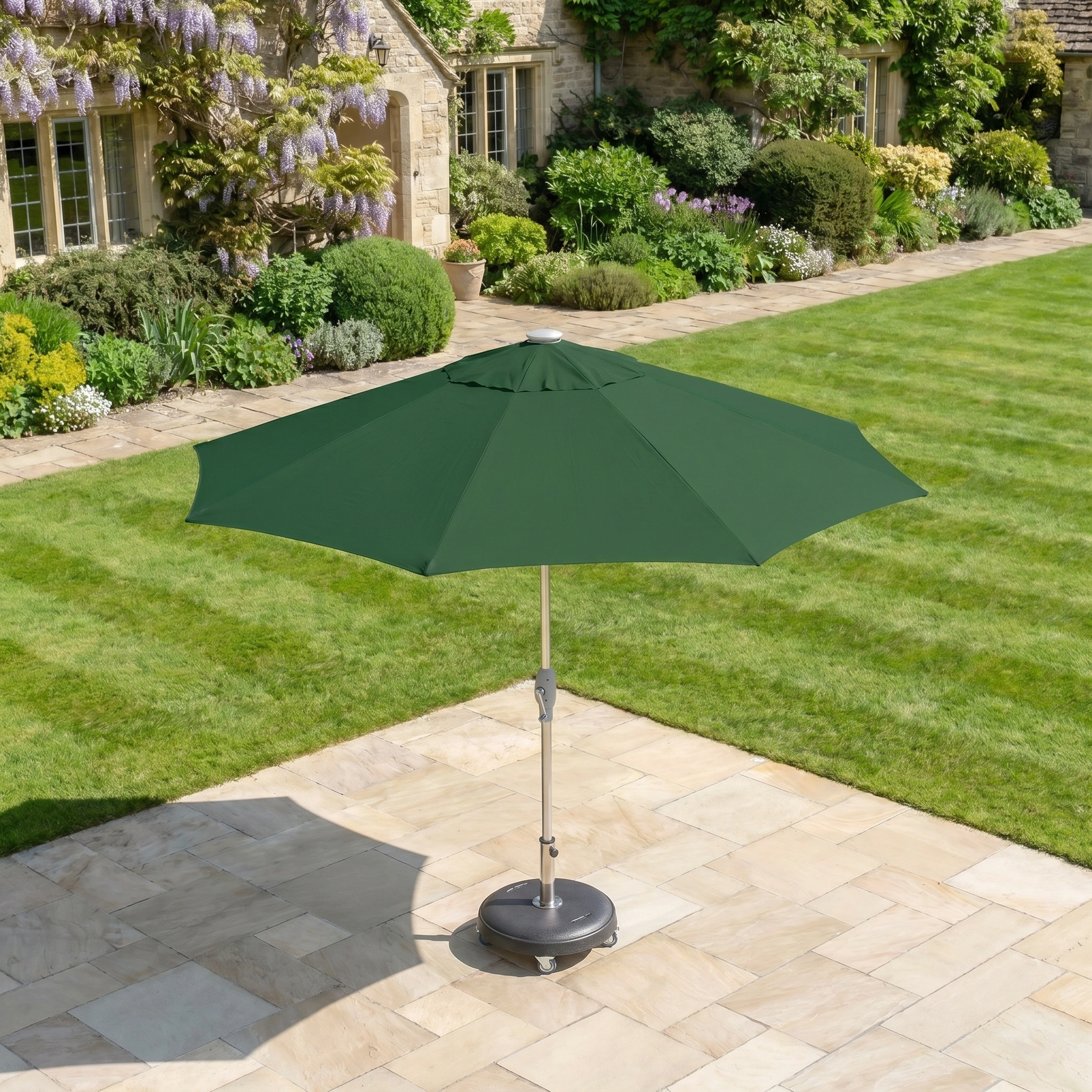 The Zelos 2.5M Round Parasol with LED Lighting in Green stands on a tiled garden area, with a lawn and a stone house visible in the background.