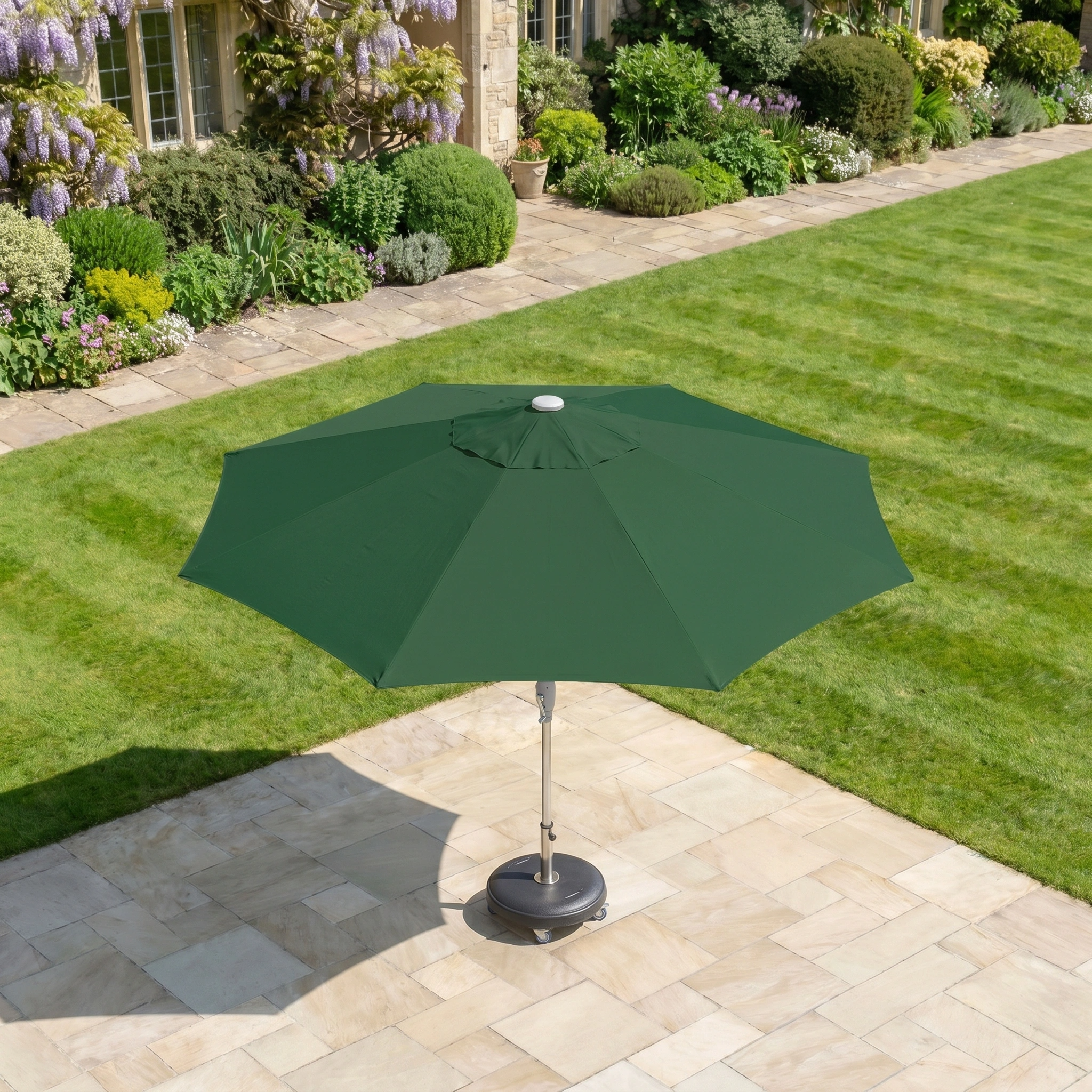 The Zelos 2.5M Round Parasol with LED Lighting in Green stands on a stone terrace beside a neatly mowed lawn and a garden filled with flowers.