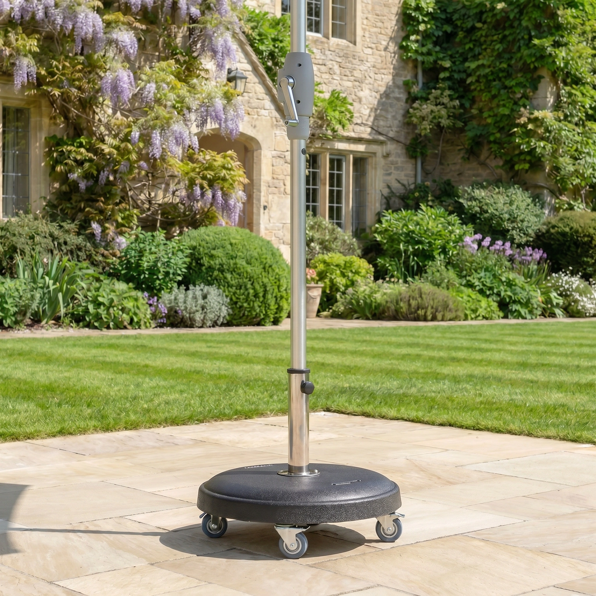 The Zelos 2.5M Round Parasol with LED Lighting in Green stands on a wheeled base atop a stone patio, surrounded by grass and blooming garden plants.