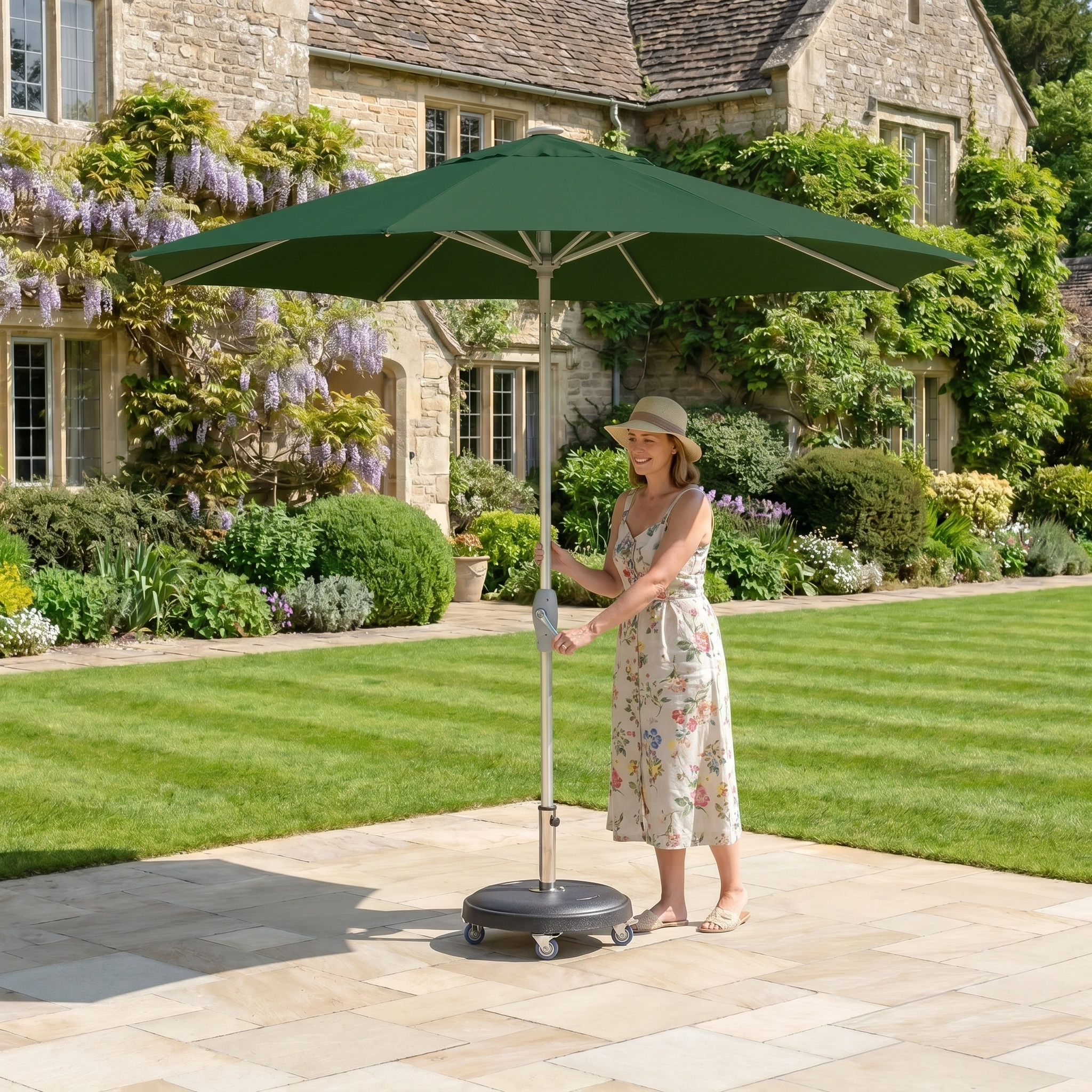 A woman in a summer dress adjusts the Zelos 2.5M Round Parasol with LED Lighting in Green in a sunny garden, with a charming stone house as the backdrop.