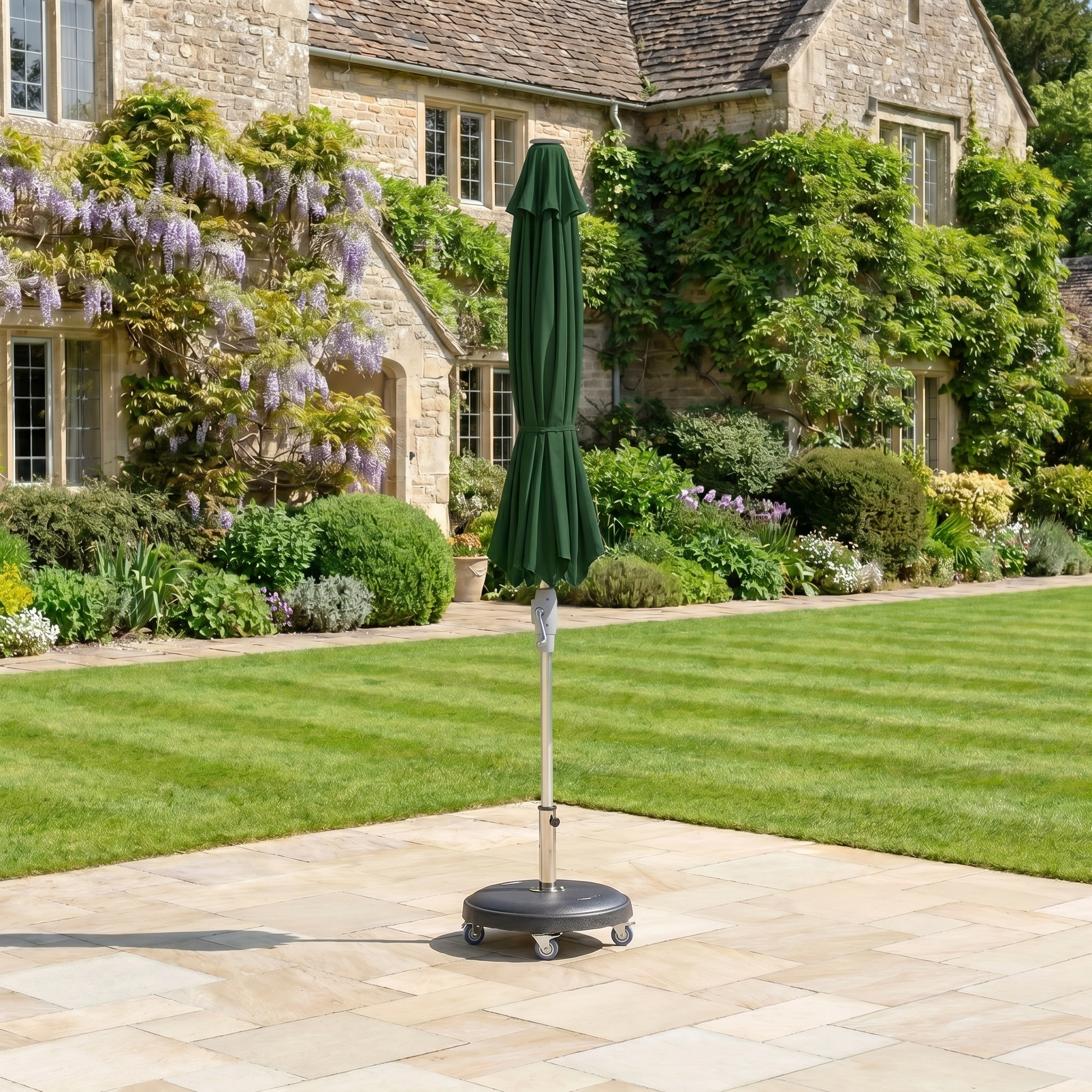 The Zelos 2.5M Round Parasol with LED Lighting in Green is closed and stands on a paved area in a garden, with a stone house visible in the background.