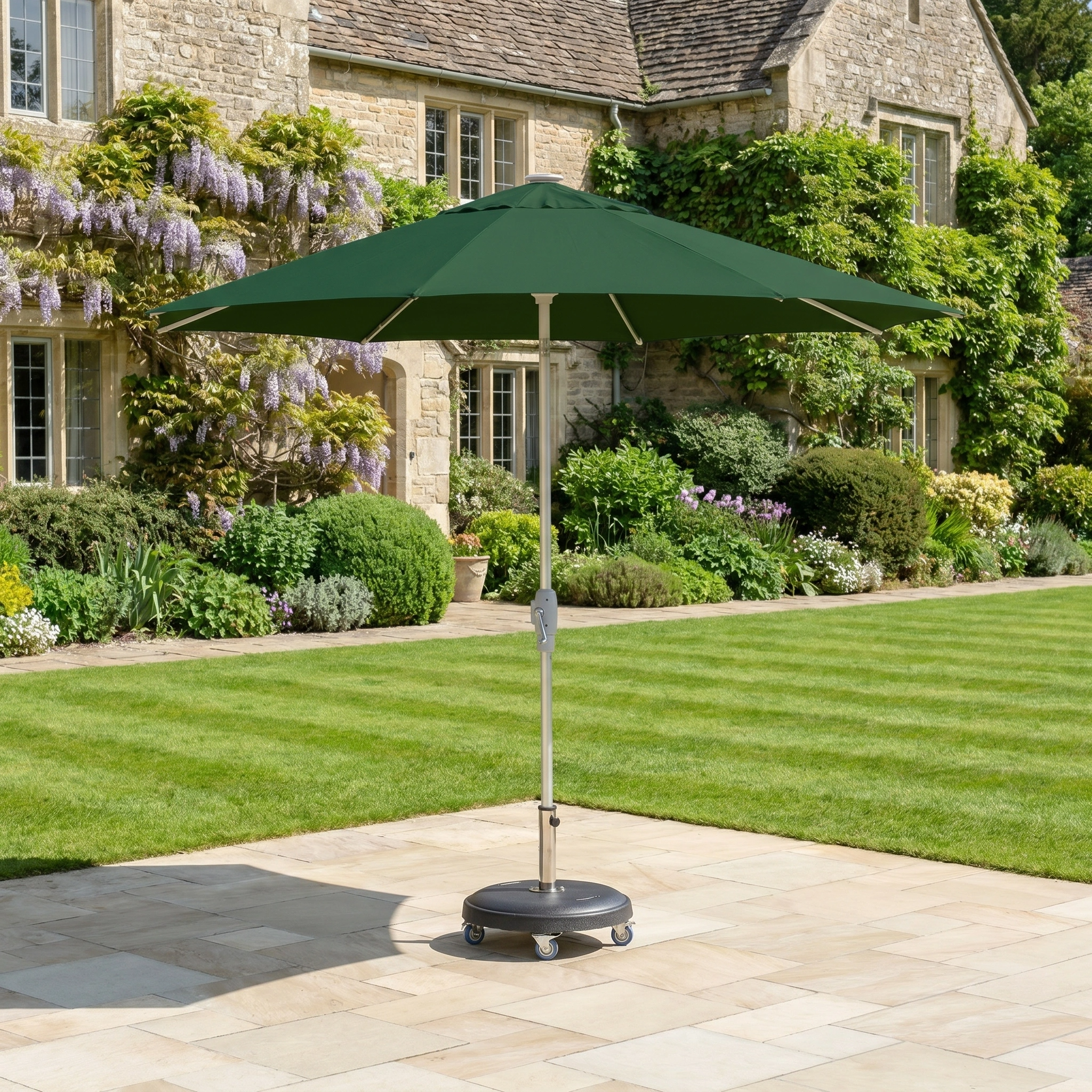 The Zelos 2.5M Round Parasol with LED Lighting in Green stands on a paved garden area, with a stone house visible in the background.