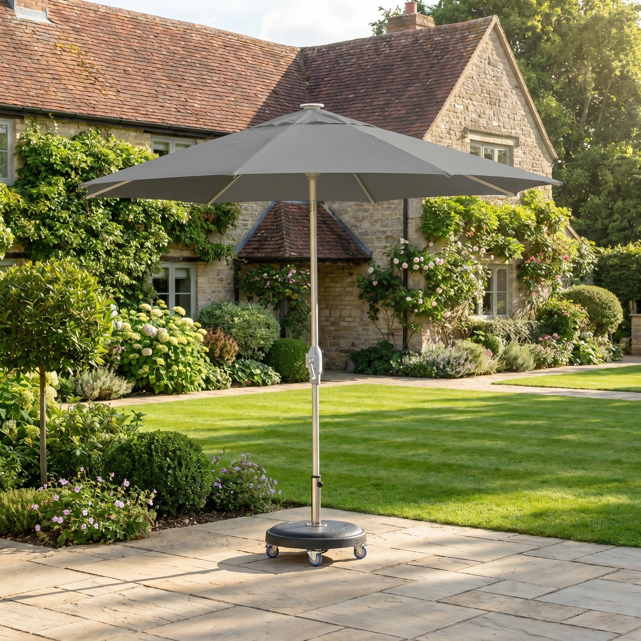 The Zelos 2.5M Round Parasol with LED Lighting in Grey stands on a stone terrace beside a stone house, surrounded by lush greenery.