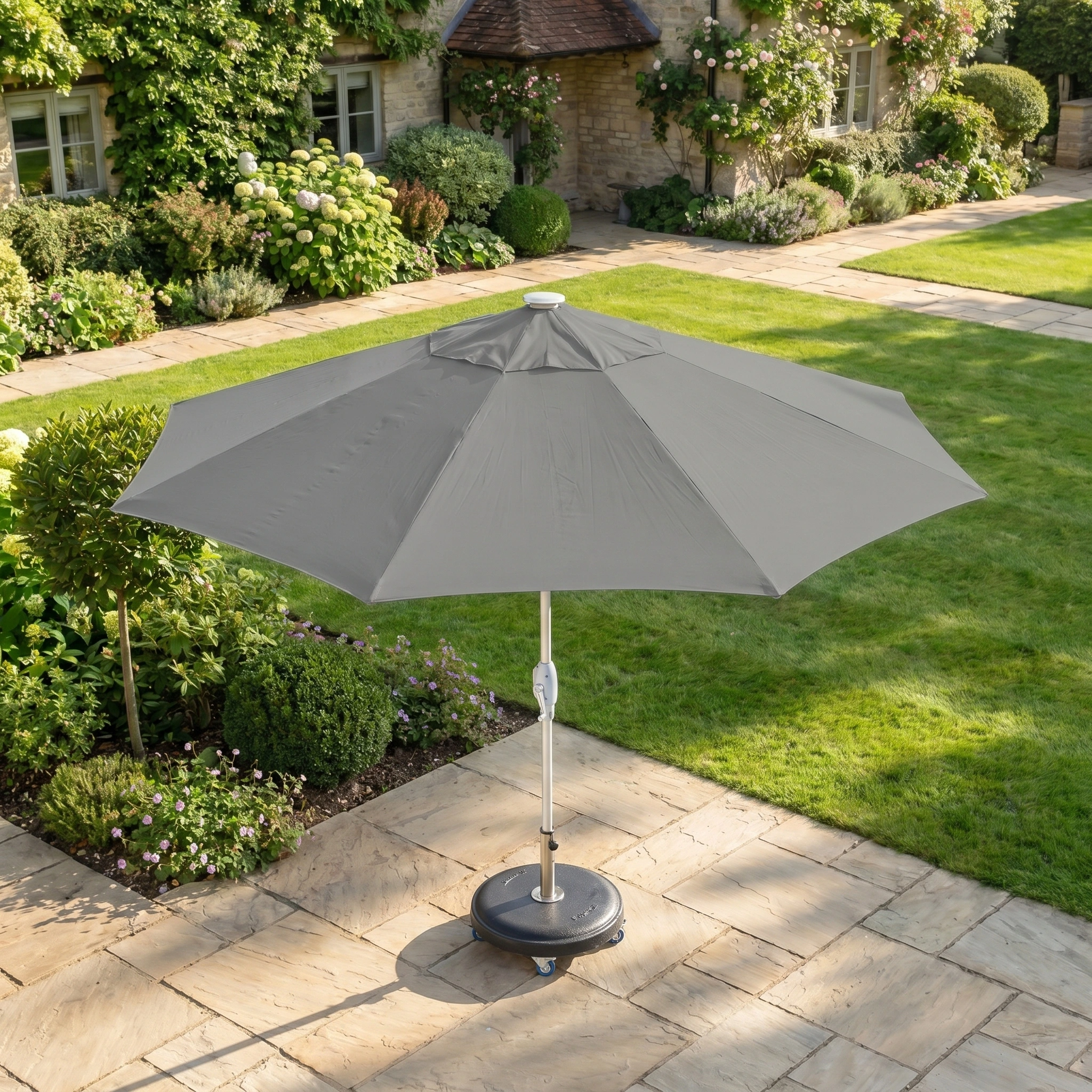 A grey Zelos 2.5M Round Parasol with LED Lighting stands open on a stone patio in a garden, offering UV protection for relaxing days outdoors amid grass and flowers.