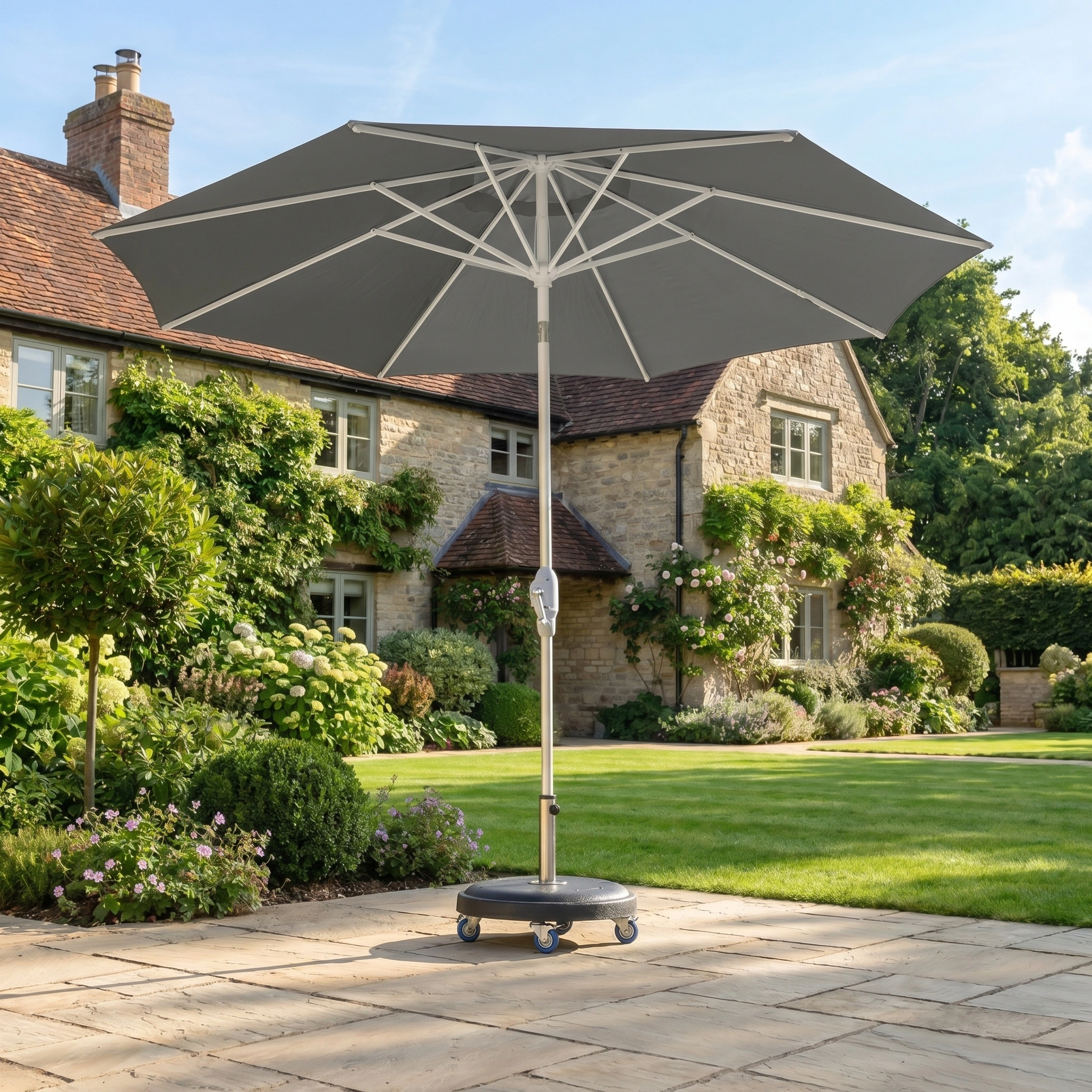 The Zelos 2.5M Round Parasol with LED Lighting in Grey stands on a stone patio by a stone house, providing UV protection and illuminated comfort for relaxing outdoors.