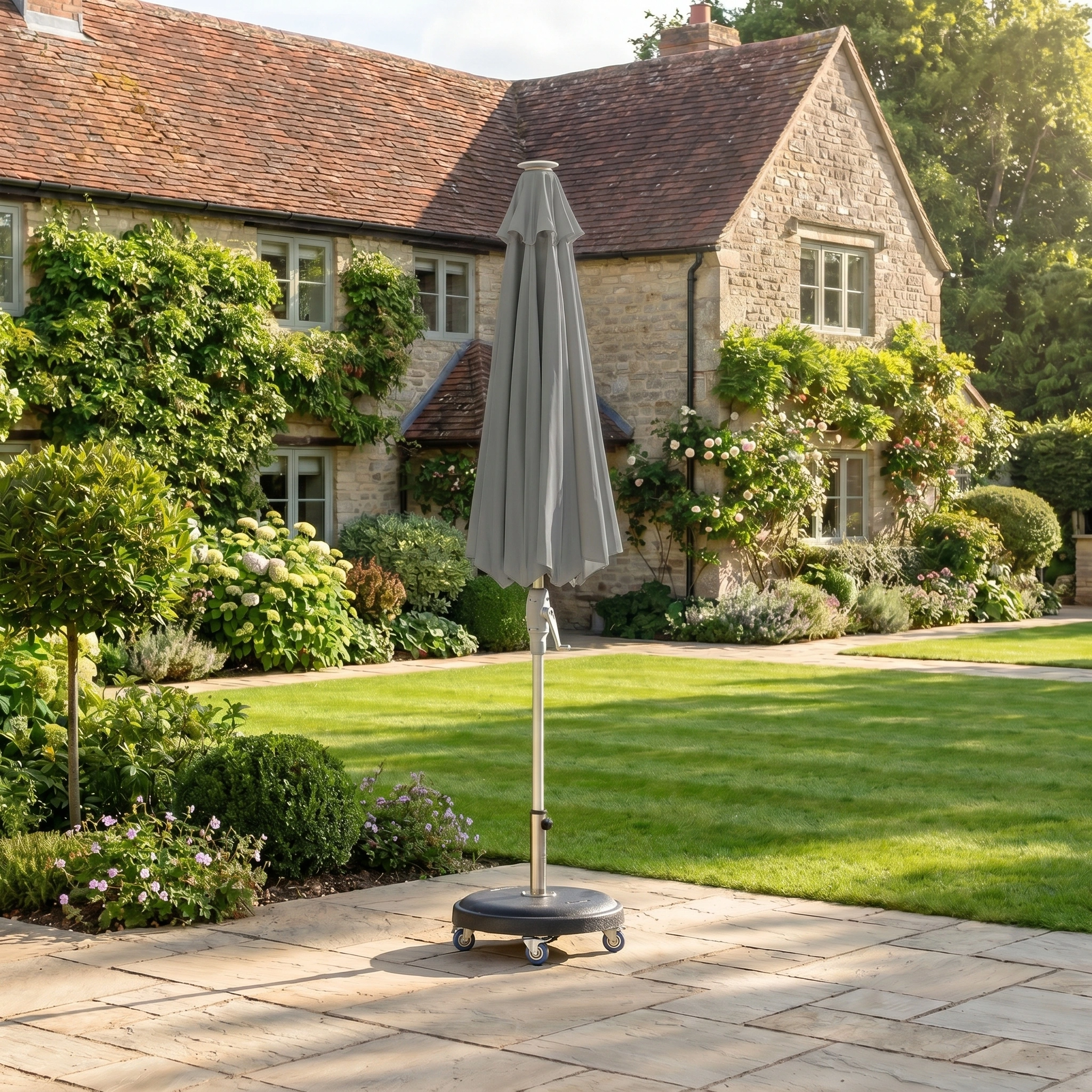 The Zelos 2.5M Round Parasol with LED Lighting in Grey stands closed on a stone terrace by a stone house with greenery, ready to provide outdoor UV protection and stylish LED lighting when opened.