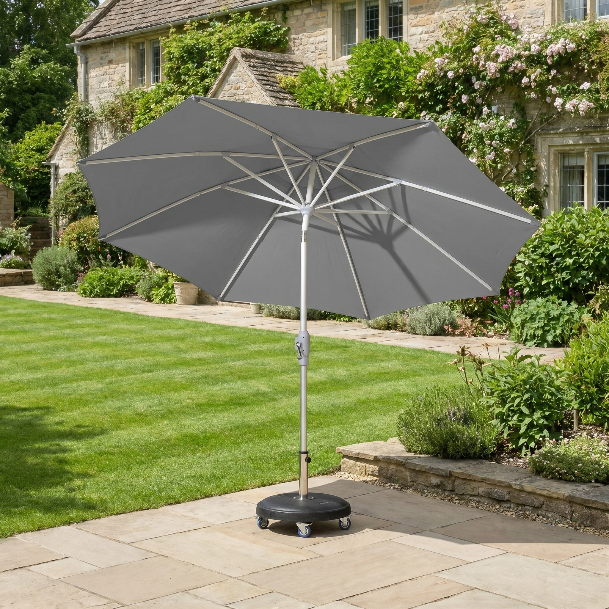 The Zelos 2.5M Round Parasol with LED Lighting in Grey stands on a black wheeled base on a paved patio by the lawn, providing stylish UV protection for outdoor spaces.