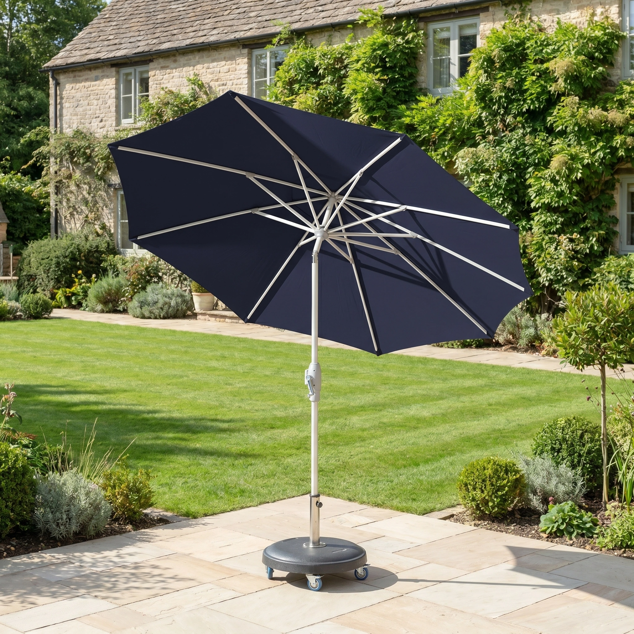 The Zelos 2.5M Round Parasol with LED Lighting in Navy stands on a stone terrace in a garden, with a house visible in the background.